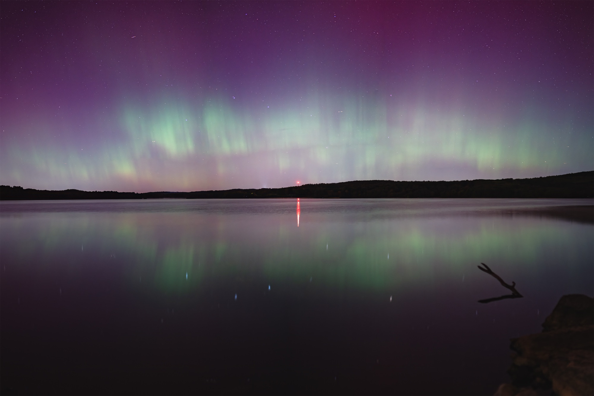 An aurora arc is reflected on a calm lake. A red radio tower is illuminated in the center of frame. 