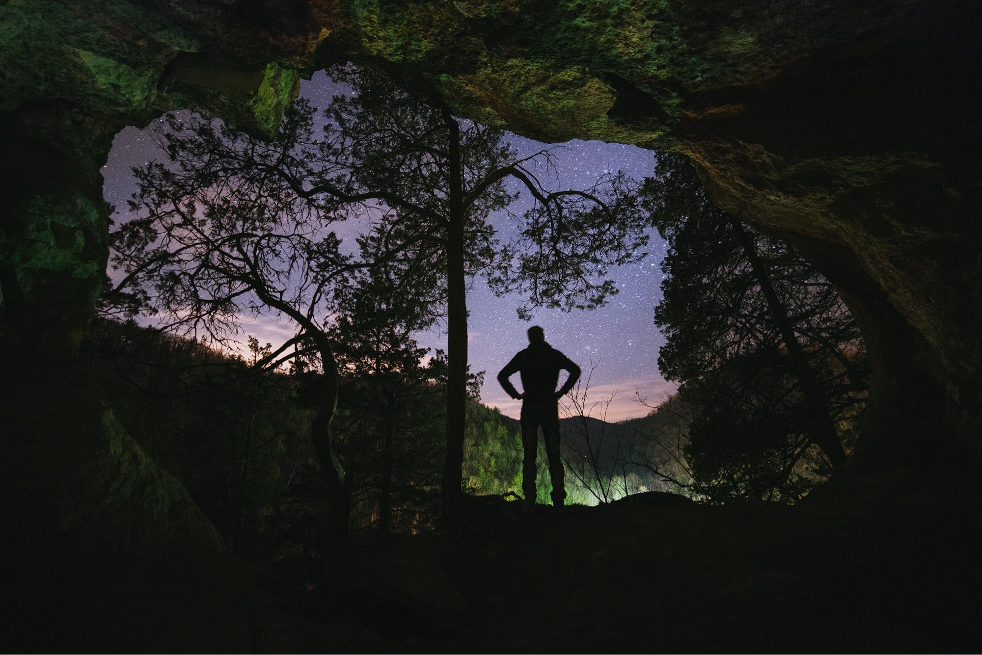 A silhouette of a man standing with arms on hips in the opening of a cave on a hill. Some nearby by trees are silhouetted against a starry night sky above some hills.