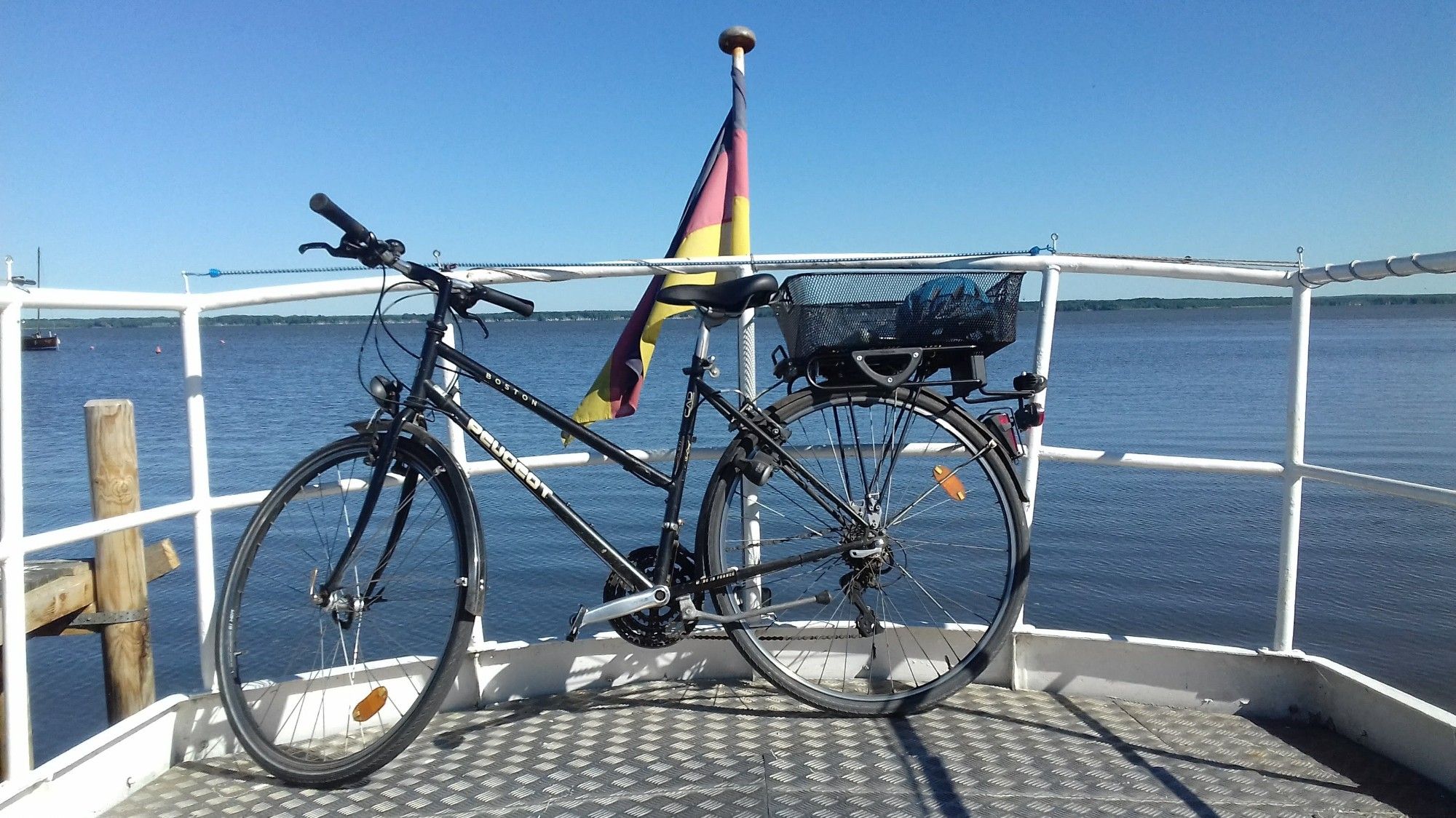 Im Bug eines kleinen weißen Schiffes steht quer vor der Reling auf dem Metallboden ein schwarzes Fahrrad. In der Ferne sieht man das bewaldete Ufer des Sees. An der Bugspitze hängt locker die Deutschlandflagge herunter.