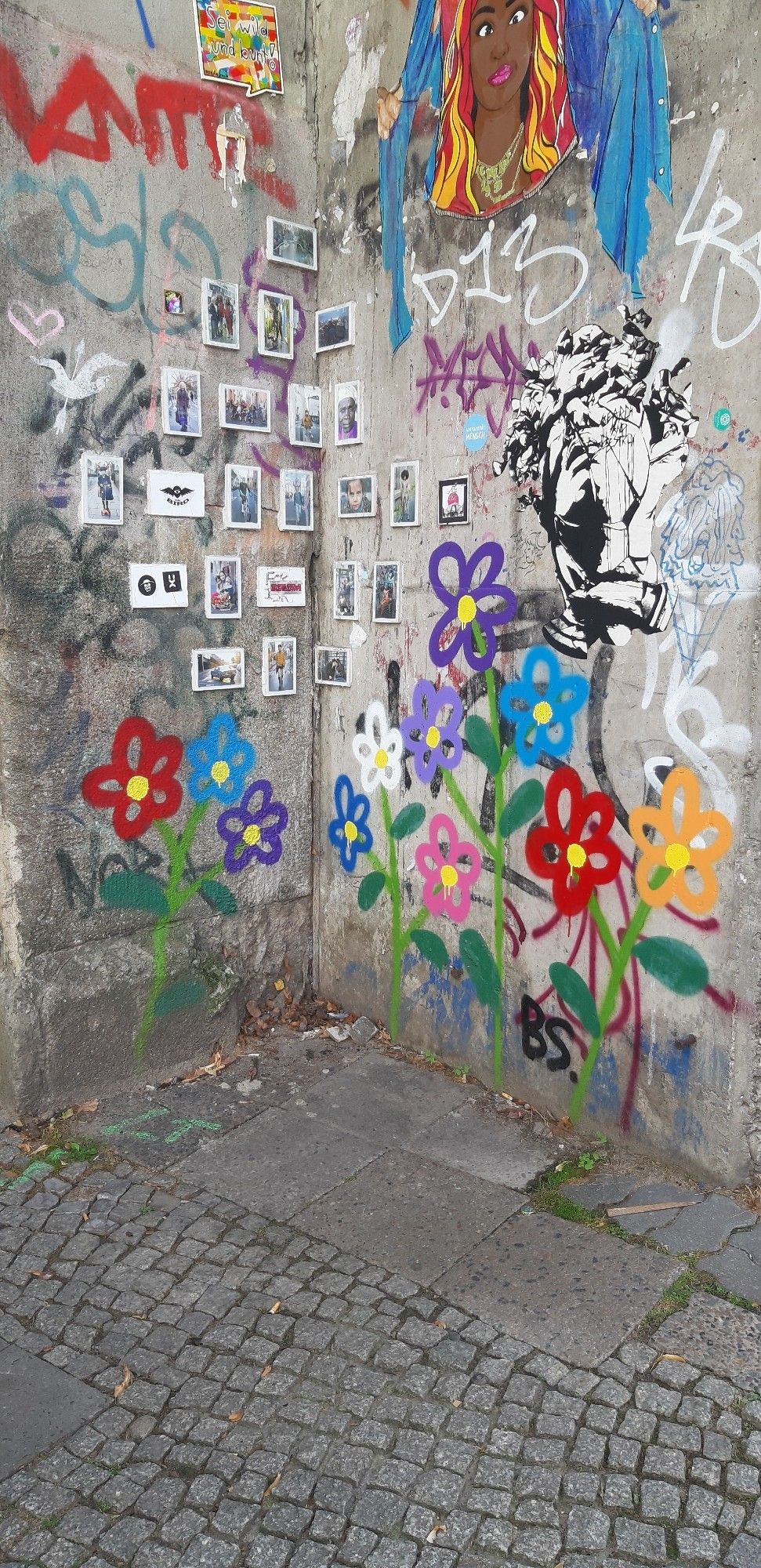 In einer grauen Hausecke hängen in Augenhöhe viele kleine gerahmte Bilder auf der veputzten Wand, meist sind Personen darauf zu sehen. Dahinter einige alte verwaschene unleserlichebSchriftzüge, darüber ein buntes Frauenportrait. Aus dem gepflasterten Boden "wachsen" schlichte große bunte Blumen auf langen grünen Stielen