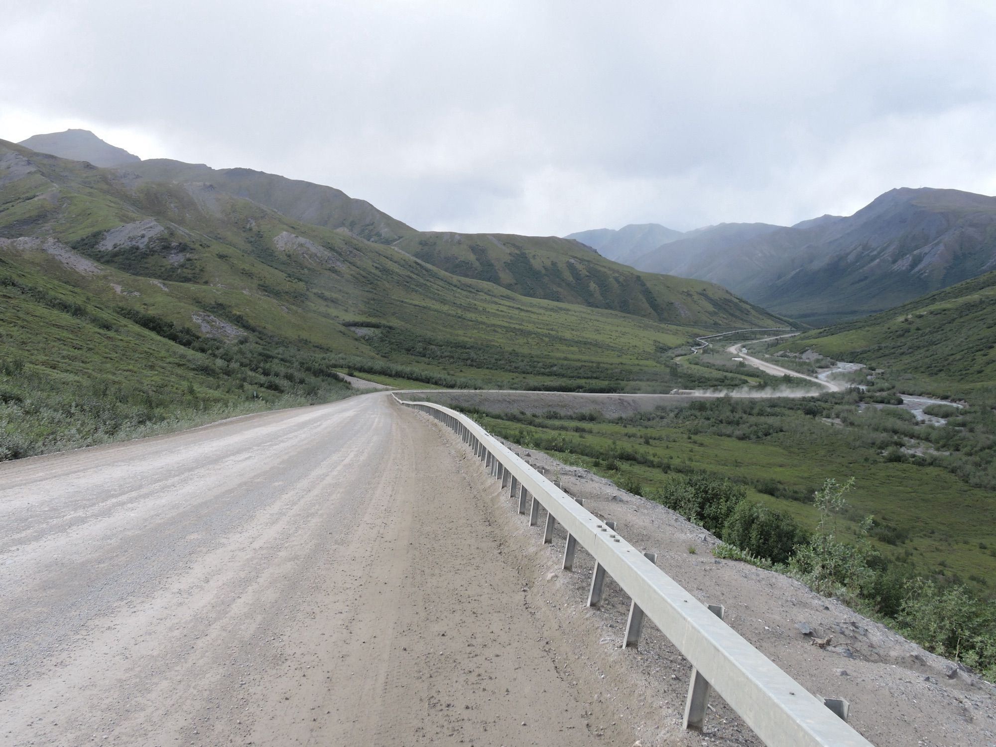 Das Bild zeigt eine kurvenreiche, unbefestigte Straße, die durch eine grüne, bergige Landschaft führt. Auf der rechten Seite ist eine Leitplanke sichtbar, die den Straßenrand sichert. Die Umgebung ist von sanften Hügeln und Bergen geprägt, die teilweise bewaldet sind. Der Himmel ist bewölkt, was auf wechselhaftes Wetter hindeutet. Ein Fluss oder Bach schlängelt sich im Tal. Links ist in der Ferne die Alaska Pipeline neben dem Highway zu sehen.