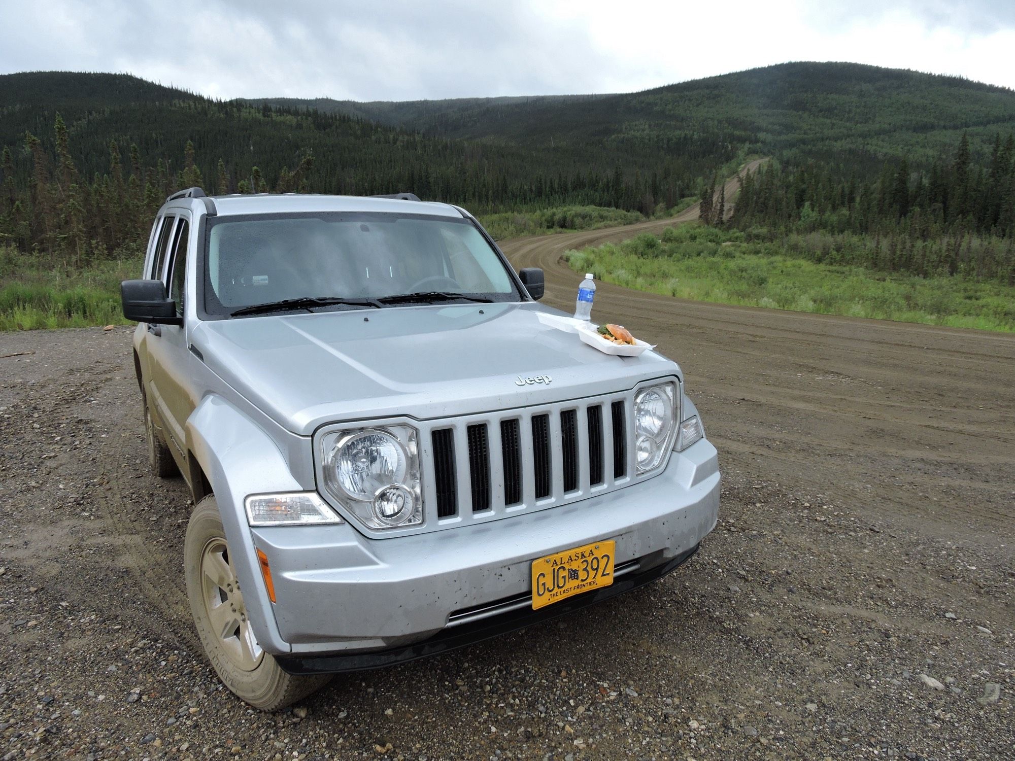 Das Bild zeigt einen silbernen Jeep, der auf einem unbefestigten Weg steht. Der Jeep hat ein Kennzeichen aus Alaska. Auf der Motorhaube liegt ein Essensteller mit einem Sandwich und einer Flasche Wasser. Im Hintergrund sind grüne Hügel und Bäume sichtbar, was auf eine ländliche oder bergige Umgebung hinweist. Der Himmel ist bewölkt, was auf mögliches wechselhaftes Wetter hindeutet.