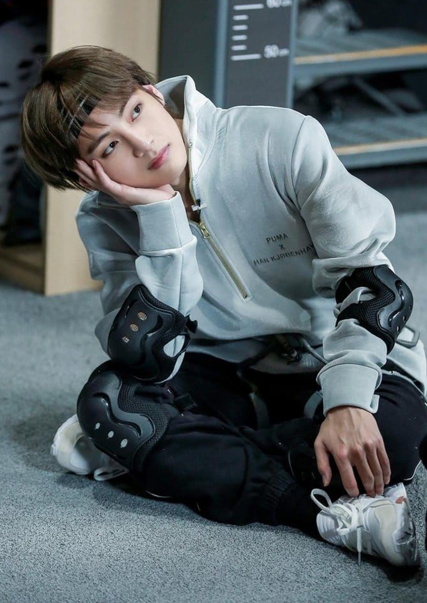 Taehyung waiting his turn to shine - gray zip up sweater, bandana around his neck, black pants, Knee and elbo guards, he is sitting cross-legged on a gray floor, head in hand, watching his brothers while he contemplates how he can beat them in the game. :)