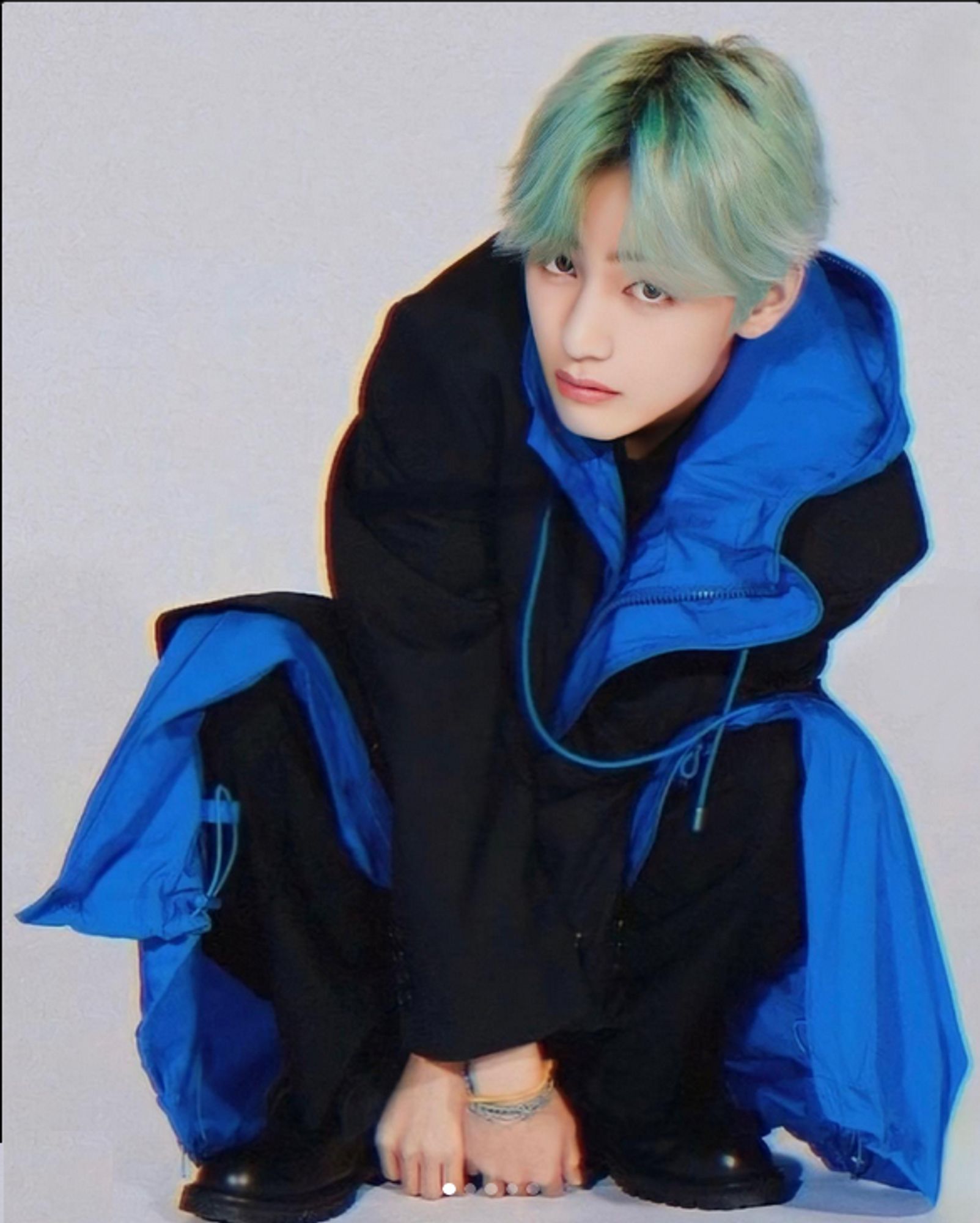 Tae in black and blue coat faded blondish-blue hair crouching with knuckles to ground, black shoes, looking at the camera, thinking who knows what.