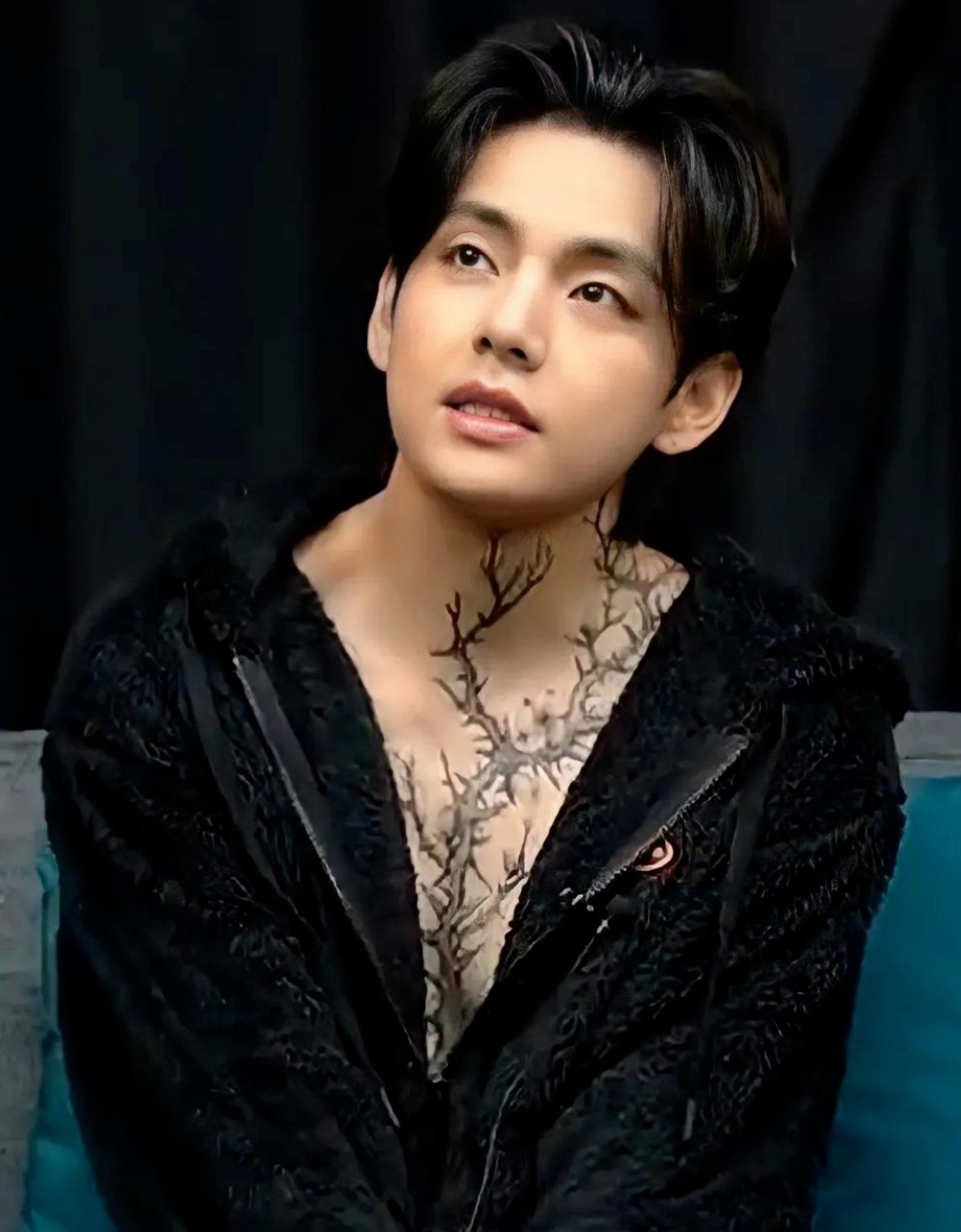 Tae on turquoise couch with pillow behind him, looking up at someone with mouth slightly open. He has a mostly unzipped fuzzy black hoodie on, showing the branch 'tattoo' in all its glory.  Wild if that had been real, yeah?