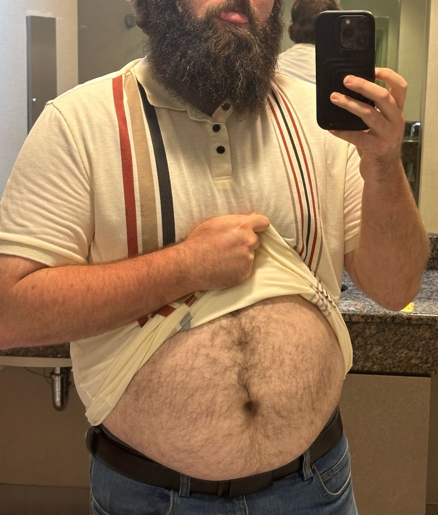 me, taking a mirror selfie, sticking my tongue out. i am lifting an off white polo shirt, with two sets of verticals stripes. thick stripes on the left which are red brown and black, and thin stripes on the right which are brown, red, black, red, brown. my belly is visible with the lifted shirt, and it is sitting on my black leather belt and jeans. you can see my belly hair, arm hair, and large beard.