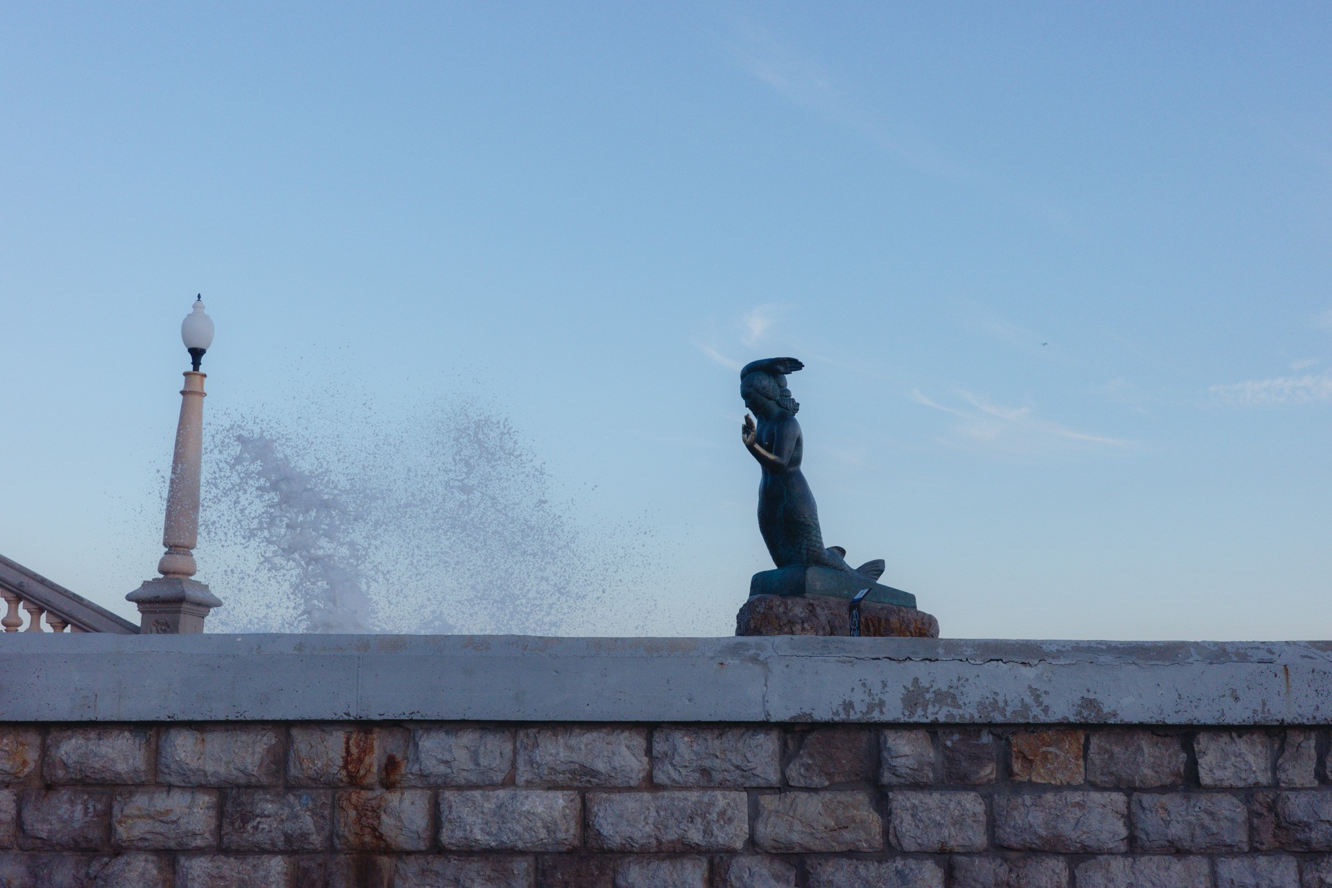 Fotografía de la estatua de la sirenita, junto a las escaleras que suben al baluarte de Sitges. Tiene una mano extendida hacia el frente, y delante de ella se elevan olas salpicando las escaleras y el paseo donde se encuentra.