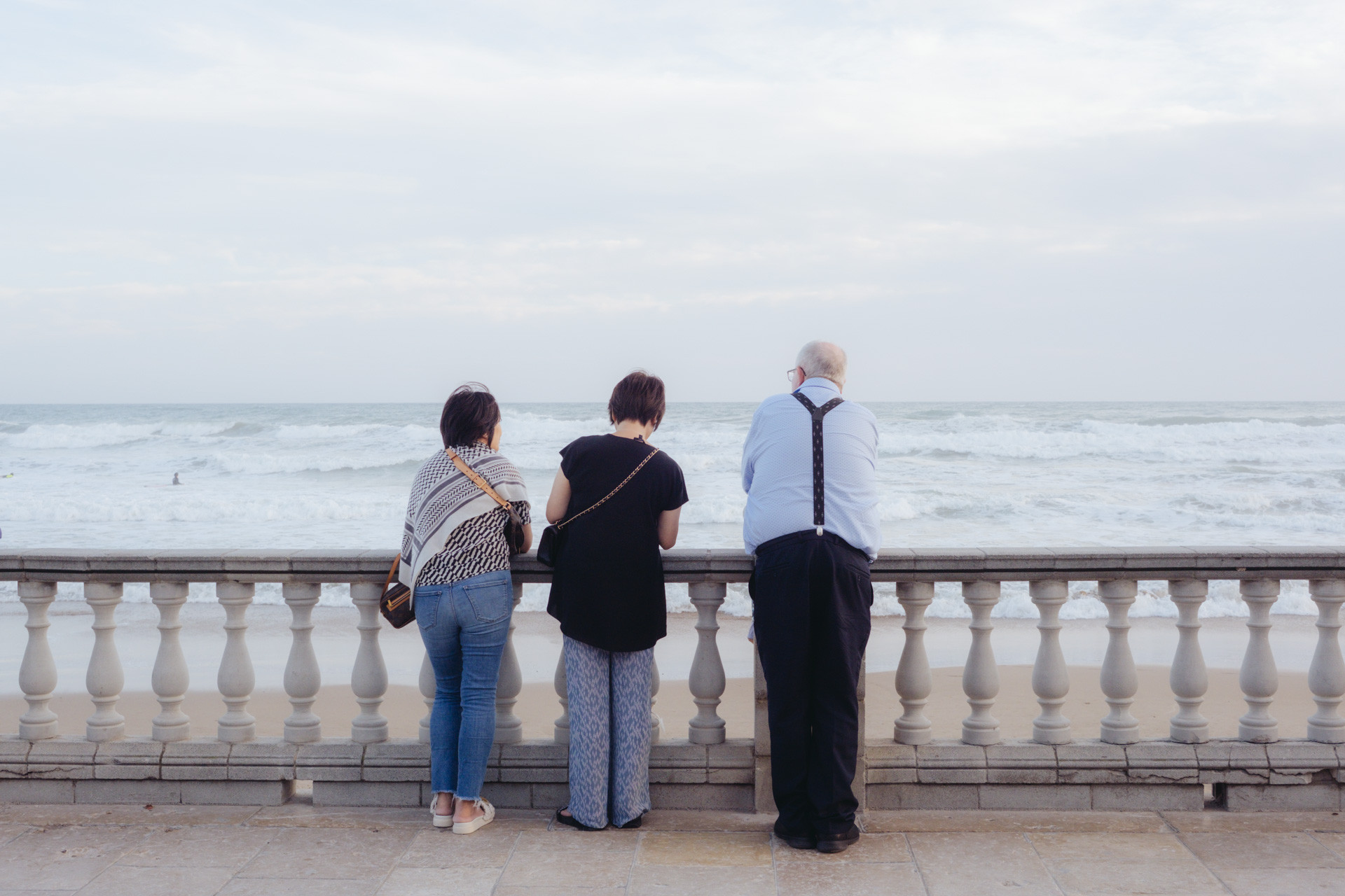 Fotografía de tres personas de espaldas mirando al mar en el paseo marítimo de Sitges. Dos son mujeres de mediana estatura, llevan el pelo corto, vestimenta similar y bolsos cruzados. El otro es un hombre considerablemente más alto y grande, viste camisa con tirantes, tiene el pelo canoso y gafas.