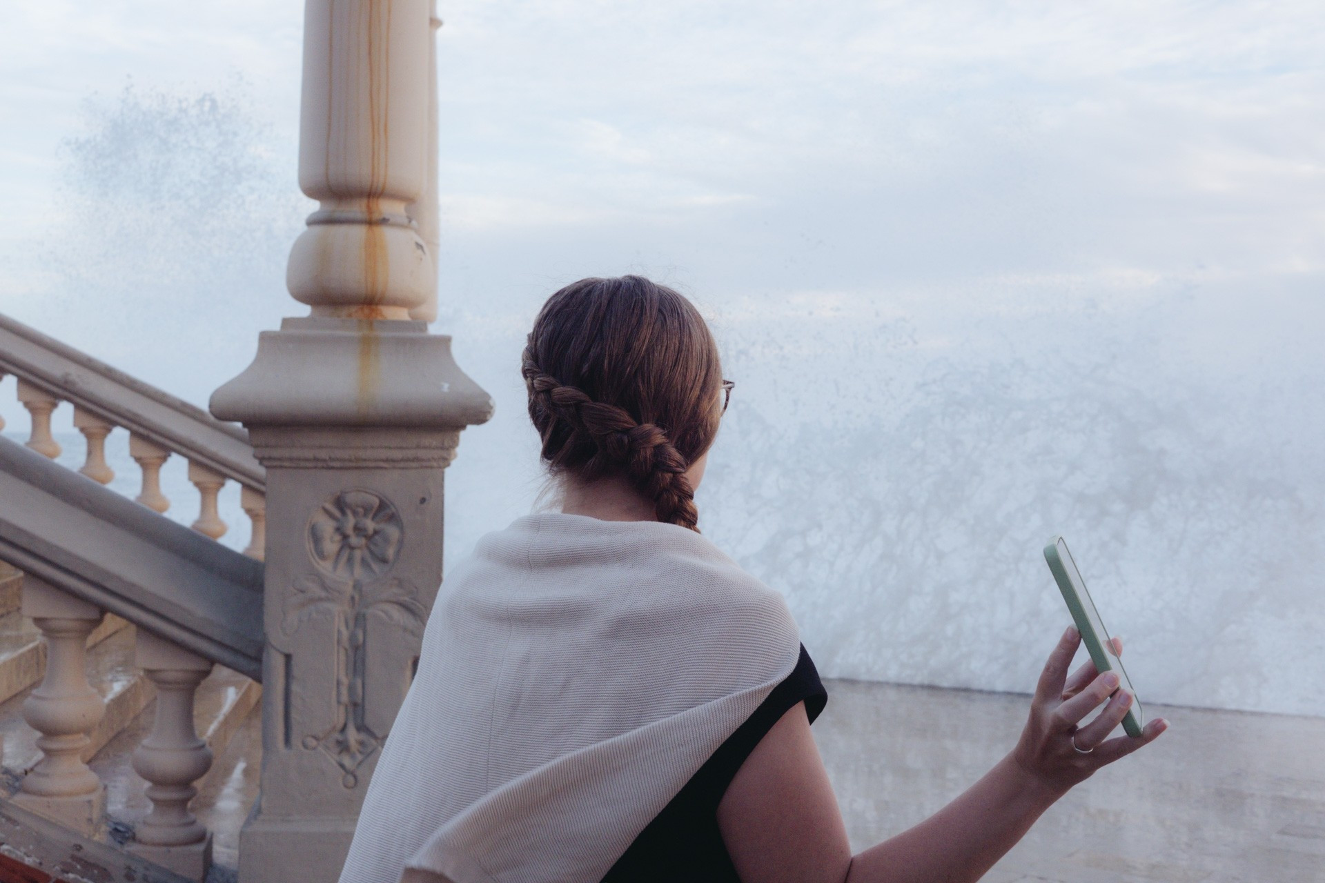 Fotografía de una mujer de espaldas, junto a las escaleras que suben al baluarte de Sitges. En la mano derecha sostiene su móvil, que le enseña a alguien que no aparece en el plano. Mira hacia el frente, y delante de ella se elevan olas salpicando las escaleras y el paseo donde se encuentra.