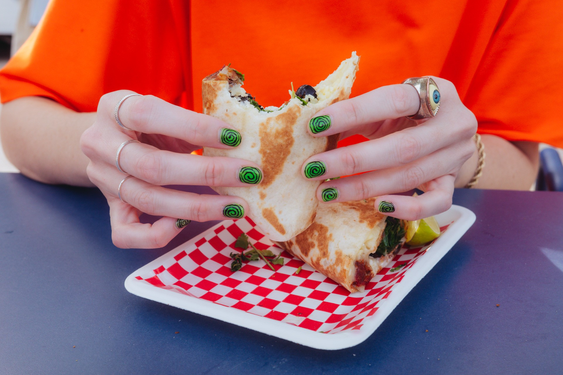 Fotografía de las manos de una mujer sosteniendo un burrito. Lleva manicura verde con espirales y un anillo con un ojo. Sostiene el burrito sobre una bandeja de cartón con un papel a cuadros rojos y blancos. La mesa donde se apoya es azul y viste una camiseta naranja. ¡Explosión de color!