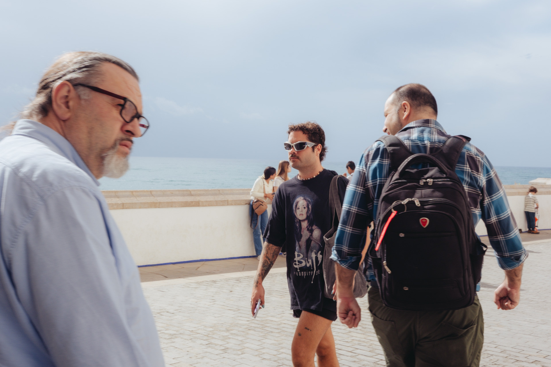 Fotografía del baluarte de Sitgess. En ella se ve a un hombre con gafas de sol, shorts y camiseta de Buffy cazavampiros andando hacia la izquierda y a dos hombres enmarcándole y dirigiéndose a la derecha. Uno le mira directamente, otro mira hacia el lado contrario. De fondo, se ve el mar.