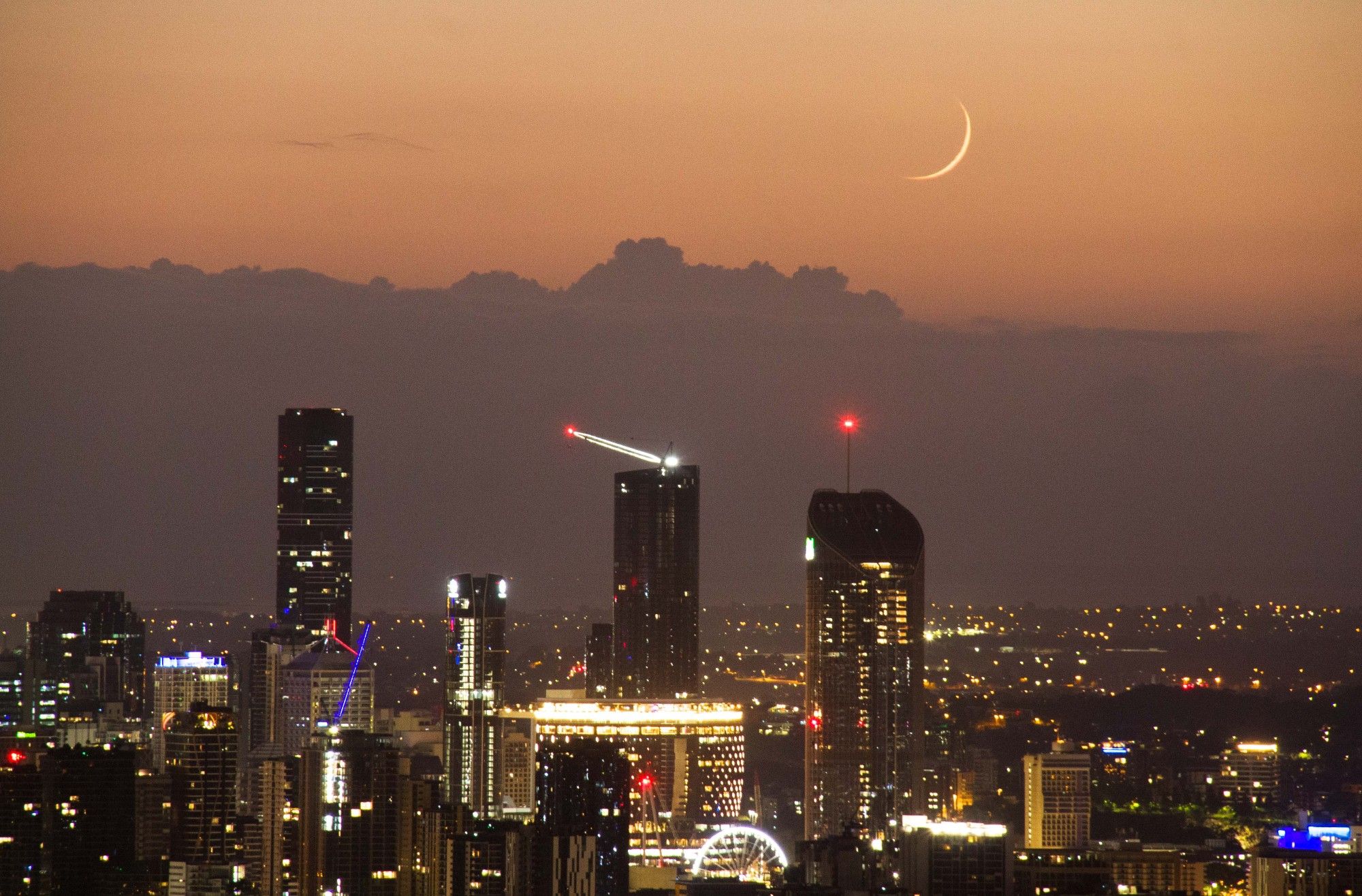 There are various towers of the Brisbane skyline, with a waning crescent moon in the background just before sunrise.