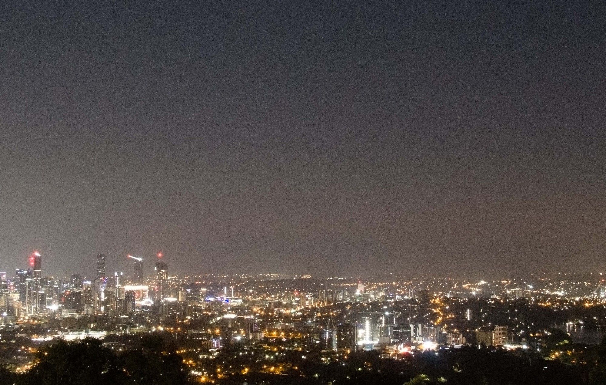 The Brisbane skyline is seen with a faintly visible comet tail nearby (to the upper right) in the background sky just before sunrise.