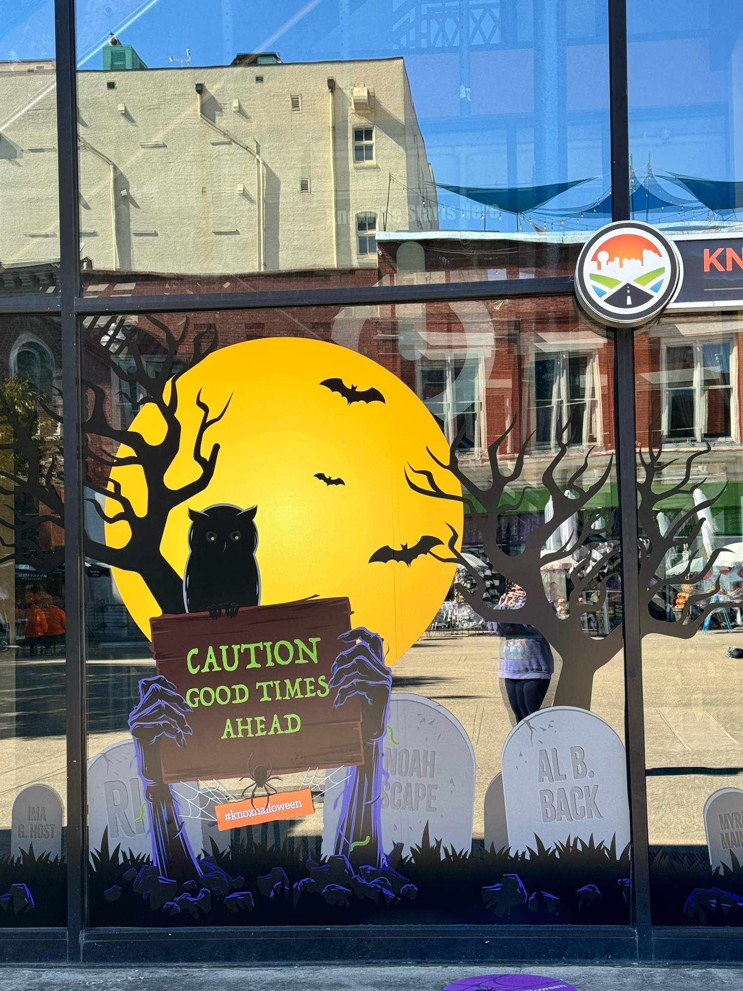 A painted window with a full moon, a black cat, some trees, some bats, and undead arms coming up from the ground holding a sign that says, “Caution Good Times Ahead.” Very Halloween-y. 