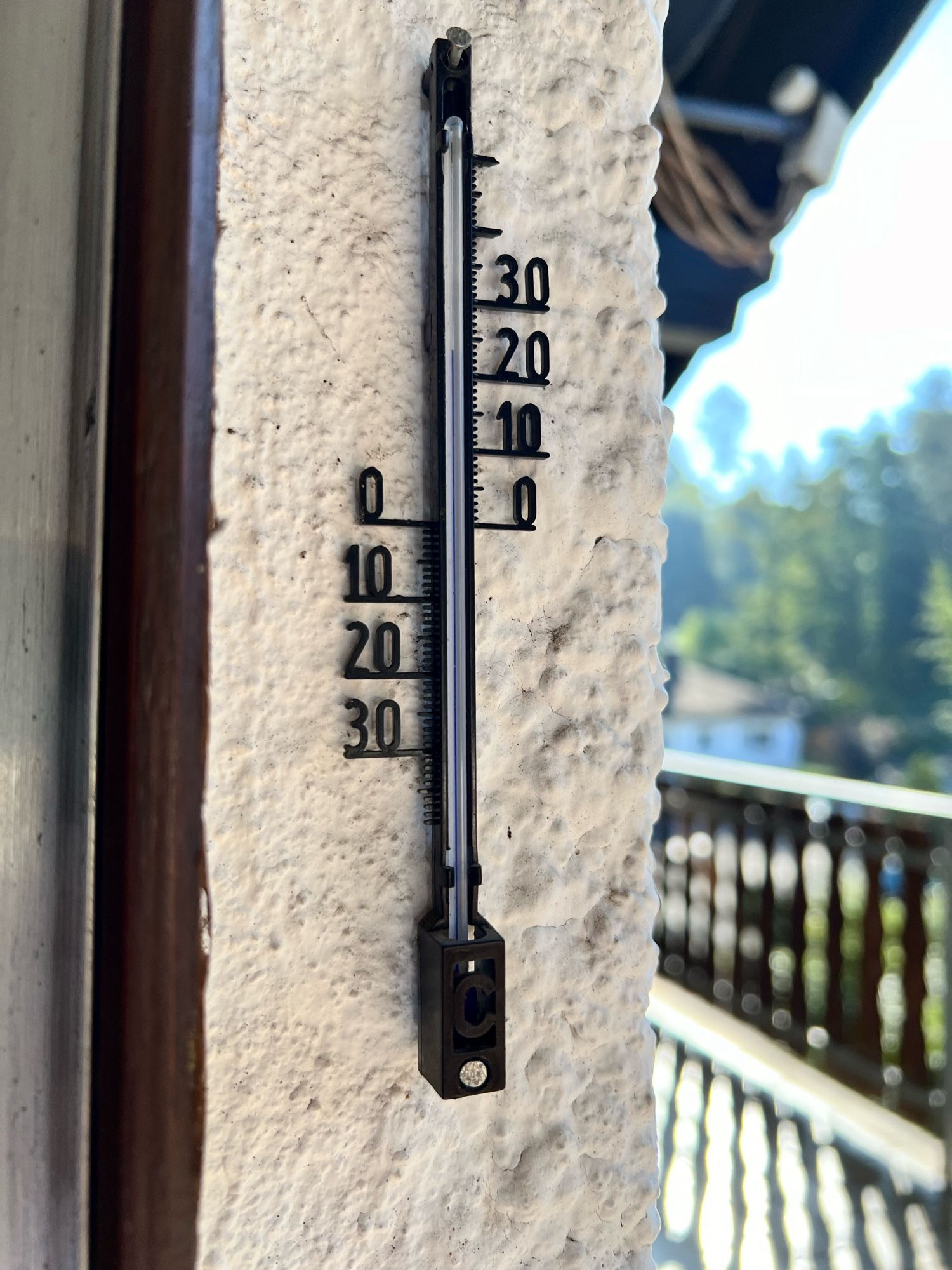 Quecksilberthermometer an der Hauswand montiert zeigt 22°. Im Hintergrund rechts ist verschwommen der Balkon und weiter dahinter der Wald zu sehen.