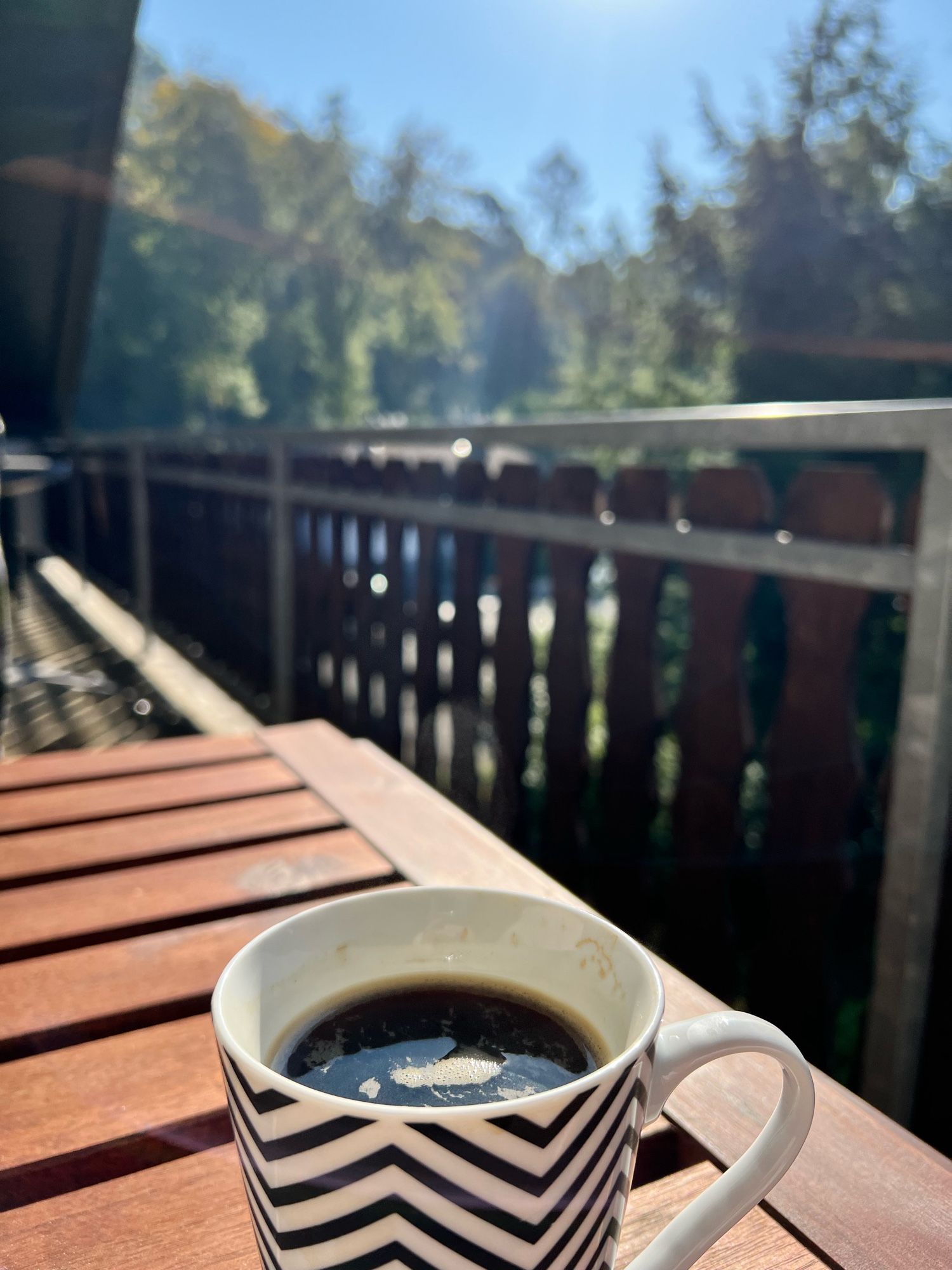 Holztisch auf dem Balkon mit einer Tasse Kaffe hinter dem Balkongeländer (rechts) der Wald, am oberen Rand blauer Himmel und ein kleines bisschen der herrlich strahlenden Sonne.