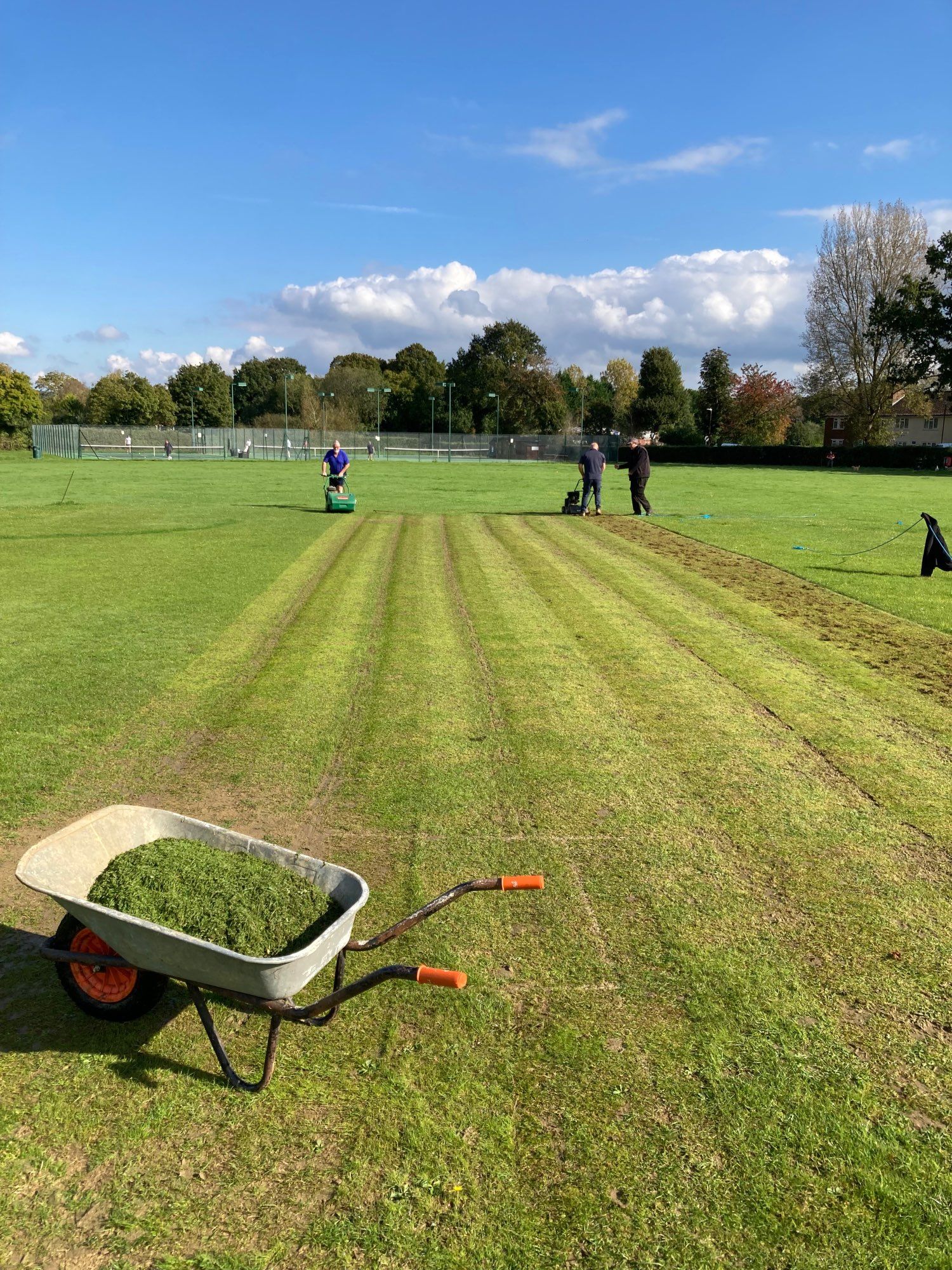 Grounds work: mowing and scarification of a cricket pitch under a blue sky.