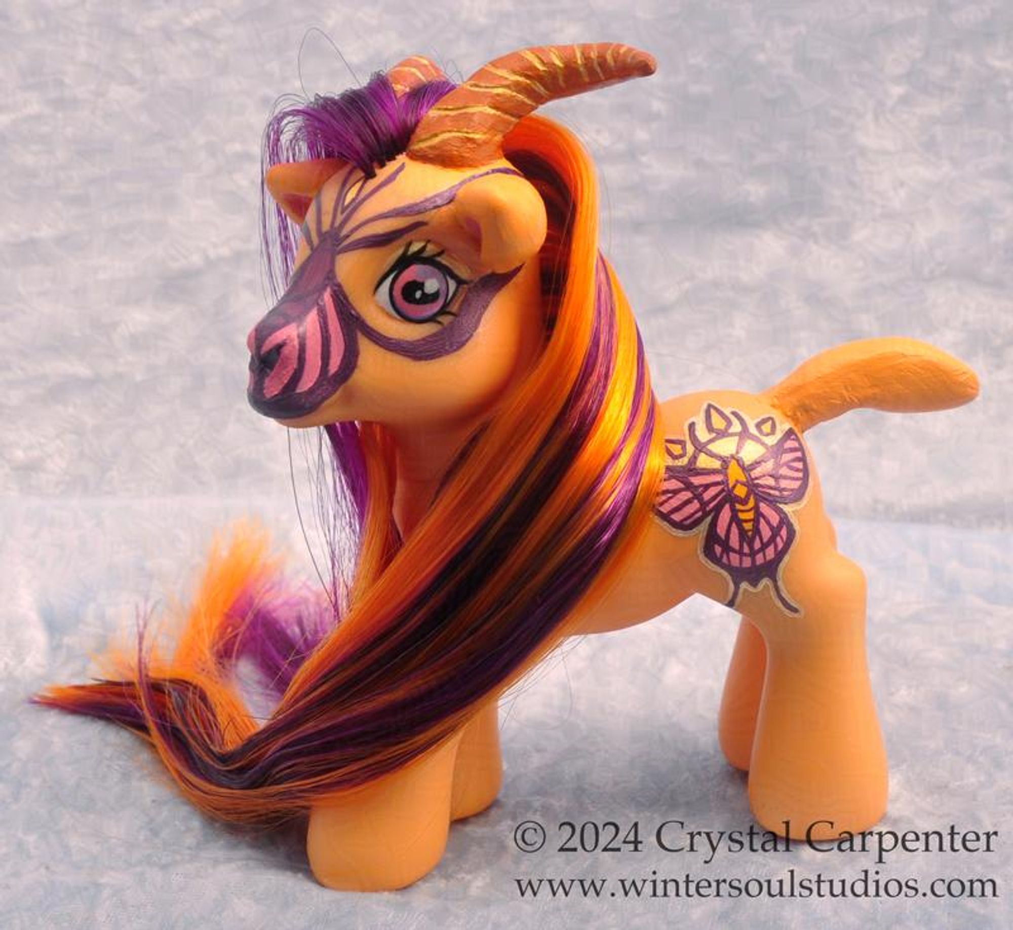 My little pony customized to be a goat. She's orange, with a gold and pink butterfly and sun cutie mark. Her long hair and beard are orange and purple. This is a clearer view of her markings.