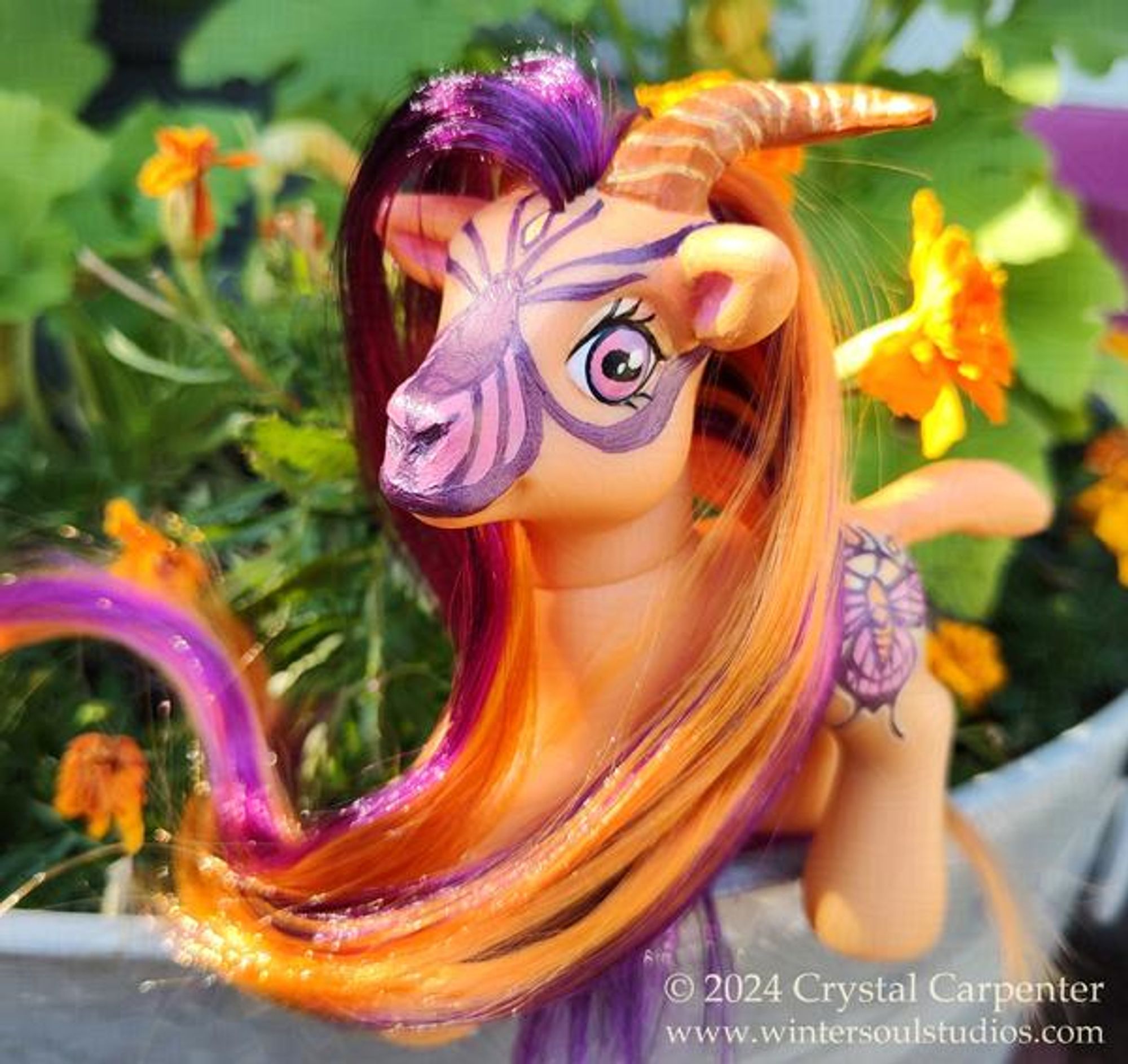 My little pony customized to be a goat. She's orange, with a gold and pink butterfly and sun cutie mark. Her long hair and beard are orange and purple. In the background are marigolds and a garden.