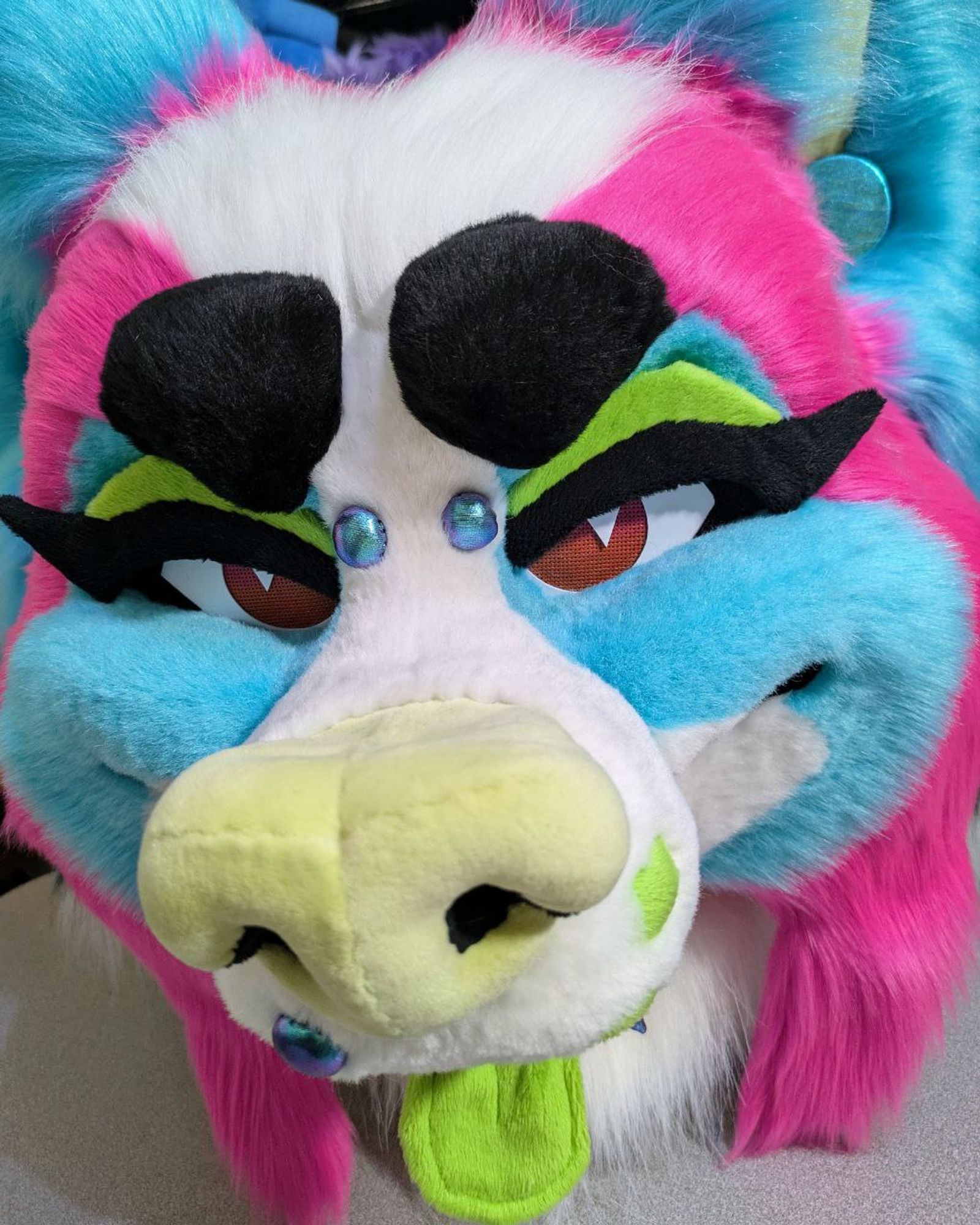 a cute, cartoony sparkledog papillon fursuit head. the fur is bright turquoise, pink, and white with green tongue, nose, and whisker dots. it has removeable green eyelids with black "cat eye" lashes and several removeable fabric "piercings" held on with magnets.