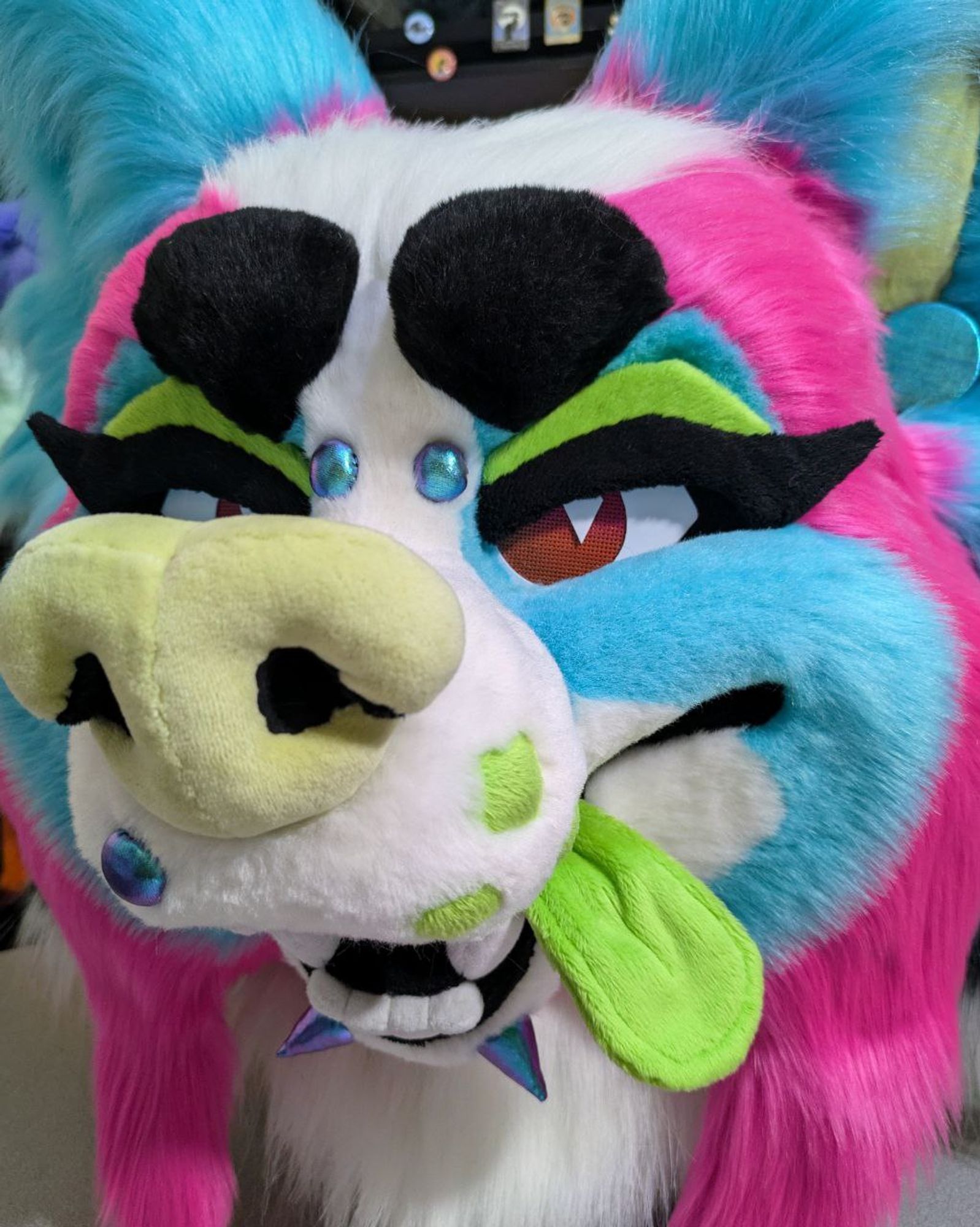 a cute, cartoony sparkledog papillon fursuit head. the fur is bright turquoise, pink, and white with green tongue, nose, and whisker dots. it has removeable green eyelids with black "cat eye" lashes and several removeable fabric "piercings" held on with magnets.