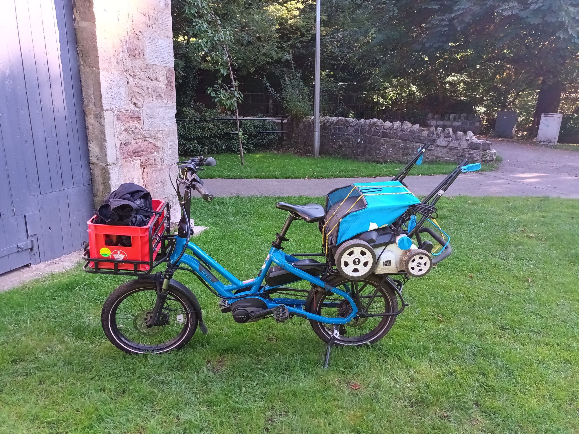 A side view of a blue Tern HSD cargo bike loaded with a lawnmower on the rear rack and a red crate of other stuff on the front rack.