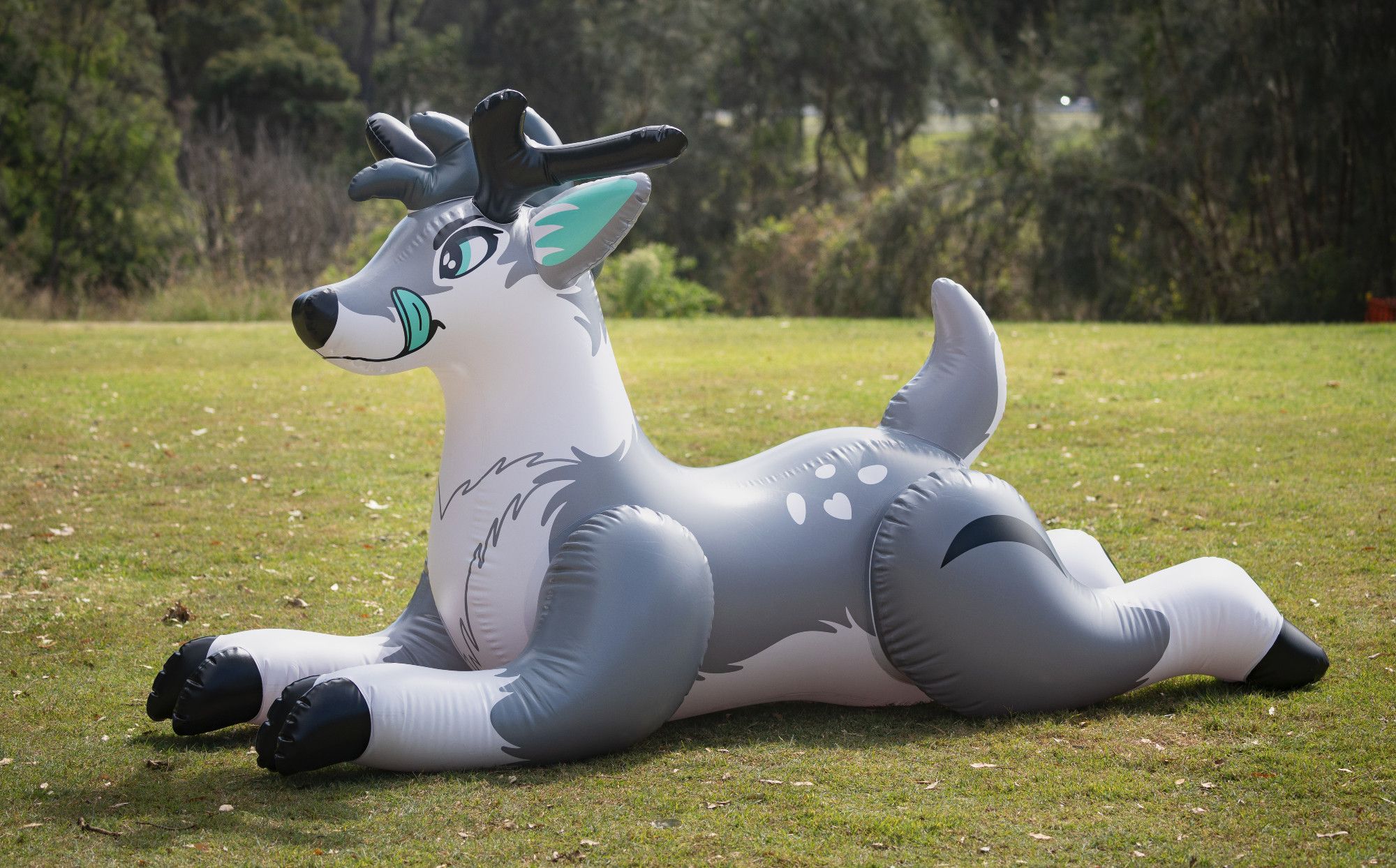 Photo of a grey inflatable deer fursona in a park surrounded by grass, with printed tongue sticking out