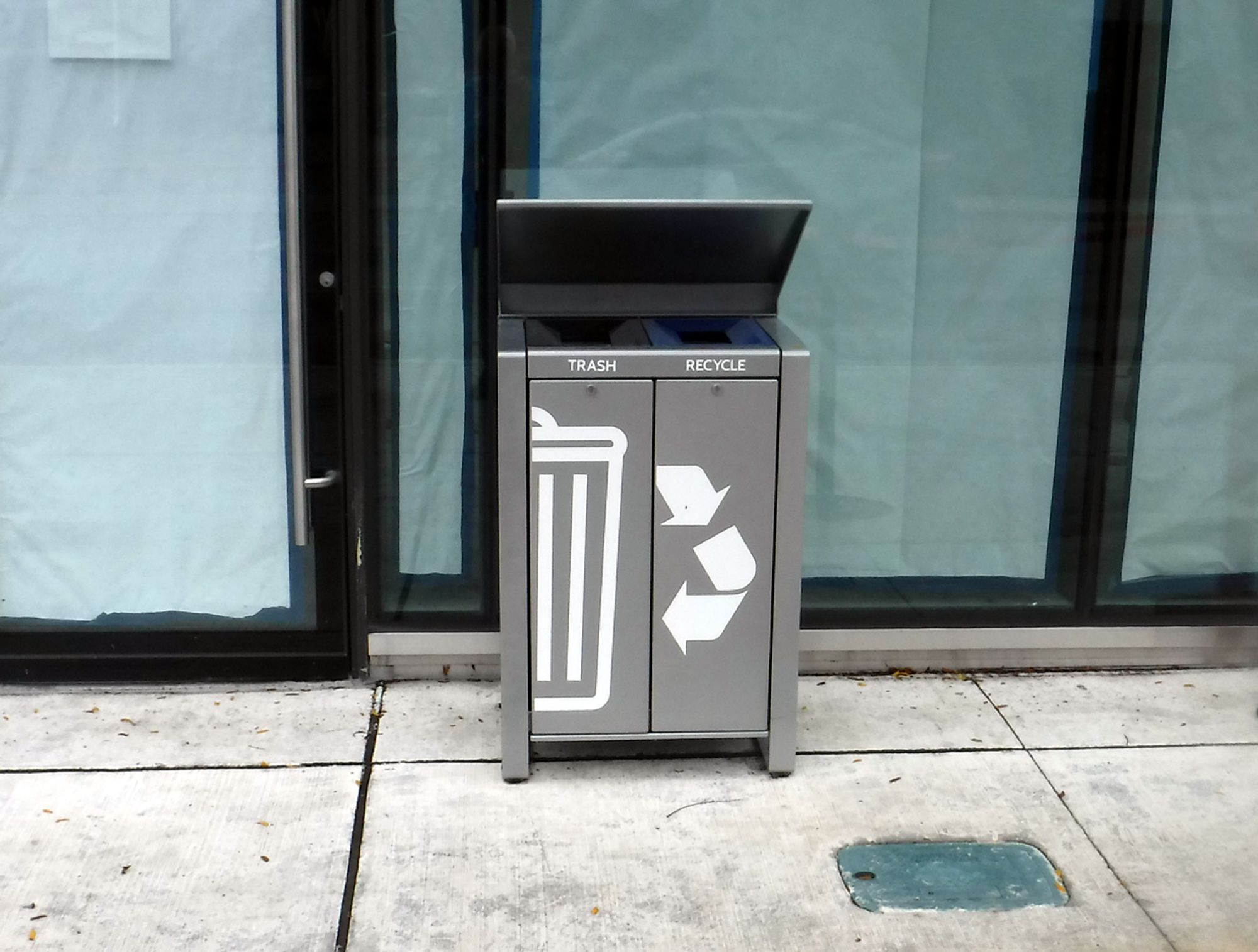 a waste bin stands in front of a new condo at 1414 Bayview Ave, with two receptacles, for Trash and Recycling, with graphics on the front showing an old-fashioned garbage can on the Trash side and stylized arrows on the Recycling side... but the arrows seem to point from the Recycling receptacle to the Trash can