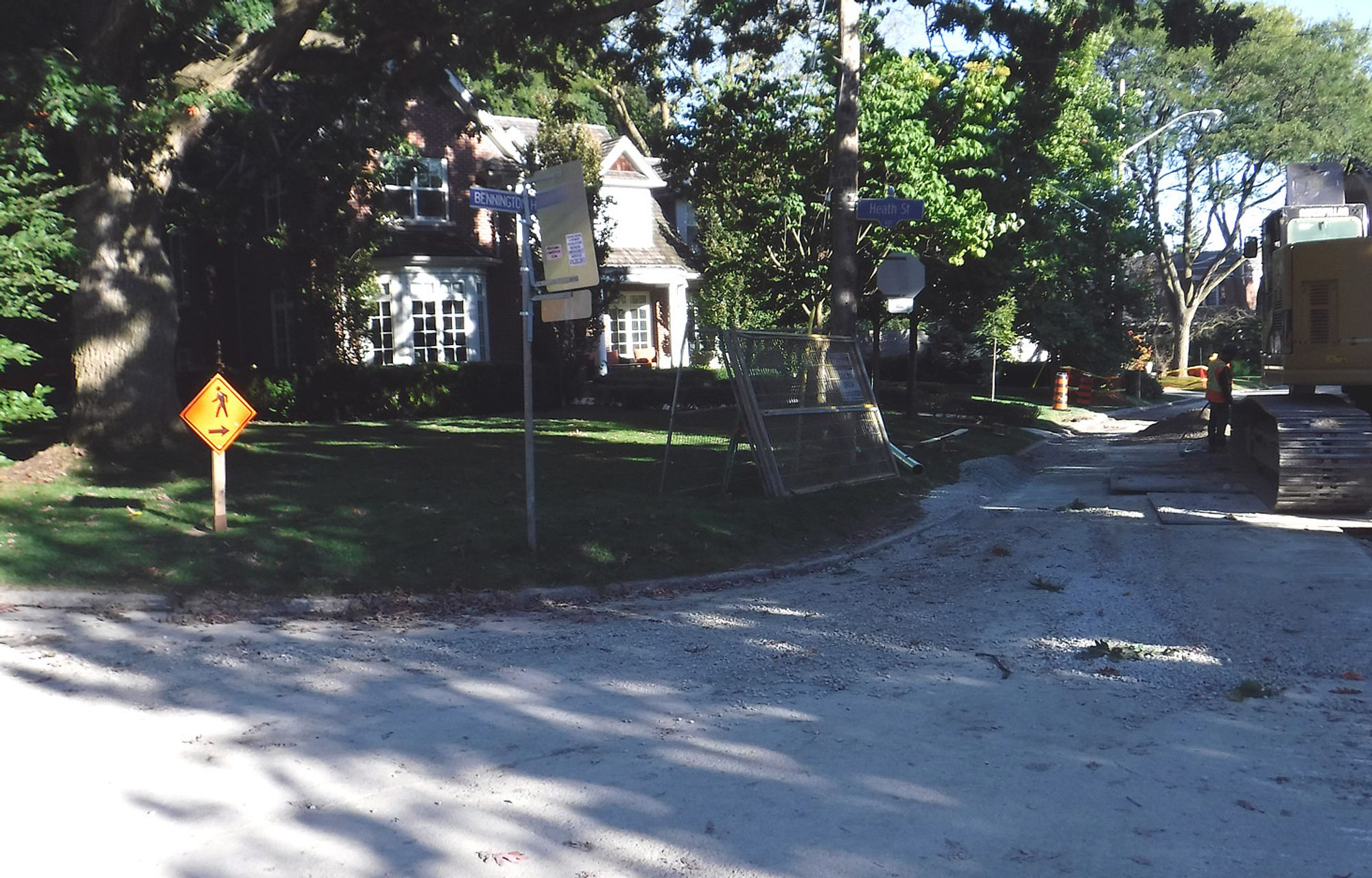 an excavator is ripping up the road surface on Bennington Heights at Heath St East... there is a yellow sign with a stylized walking pedestrian, and an arrow pointing in the opposite direction