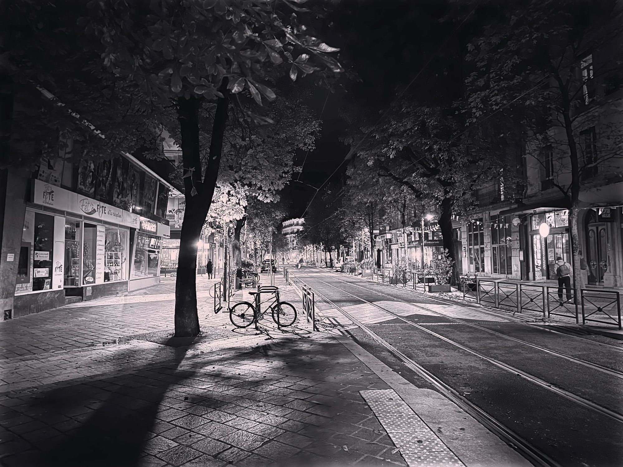 Black and white photo of a pedestrian street (with tram tracks in the middle) in Grenoble, France, late in the night.  There’s a cluster of street lights in the distance, and a single bike is hanging out on a bike rack.