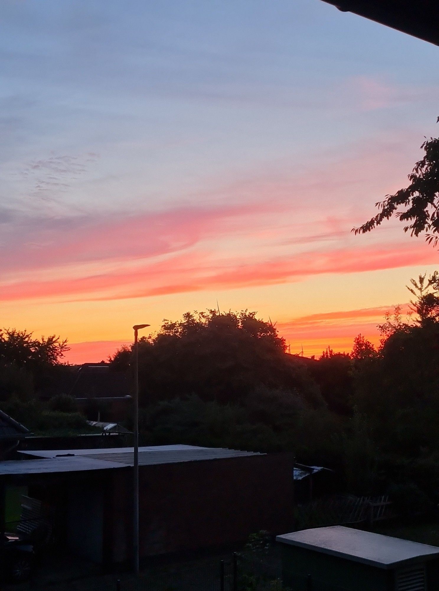 Das Bild zeigt einen wunderschönen Sonnenuntergang mit intensiven Farbtönen, die von Gelb über Orange bis hin zu einem leuchtenden Rosa und Violett reichen. Der Himmel ist teilweise von Wolken durchzogen, die die Farben des Sonnenuntergangs widerspiegeln. Im Vordergrund sind dunkle Silhouetten von Bäumen, Gebäuden und einem Lichtmast zu sehen, die den Blick auf den farbenfrohen Himmel freigeben. Die Szene strahlt Ruhe und Schönheit aus und fängt die friedliche Atmosphäre des Abends ein.