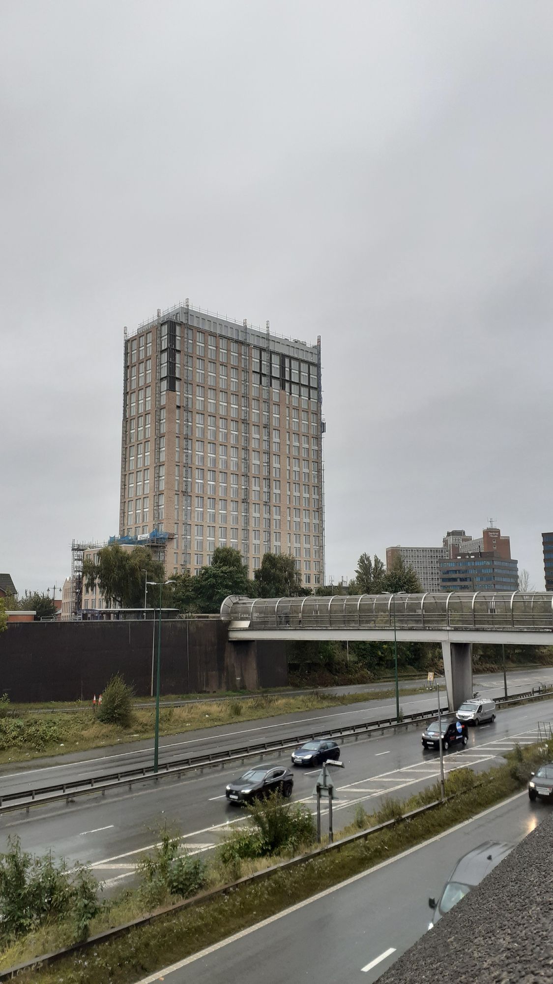 The building against a grey sky. The road surface is wet. The brick cladding has reached the top on one side, and half of the side facing us.