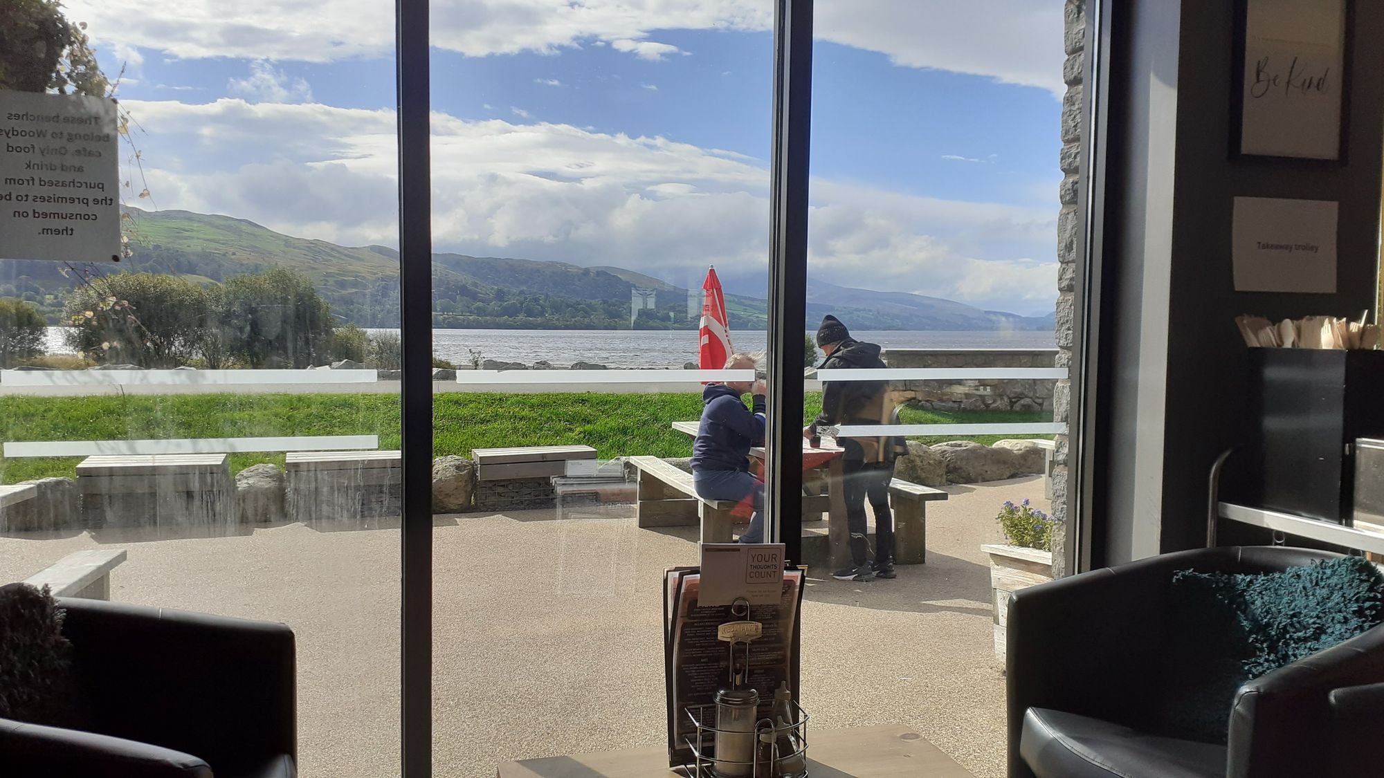 A view through cafe windows of a lake with mountains behind. It is sunny, but the tops of the mountains are in cloud. 