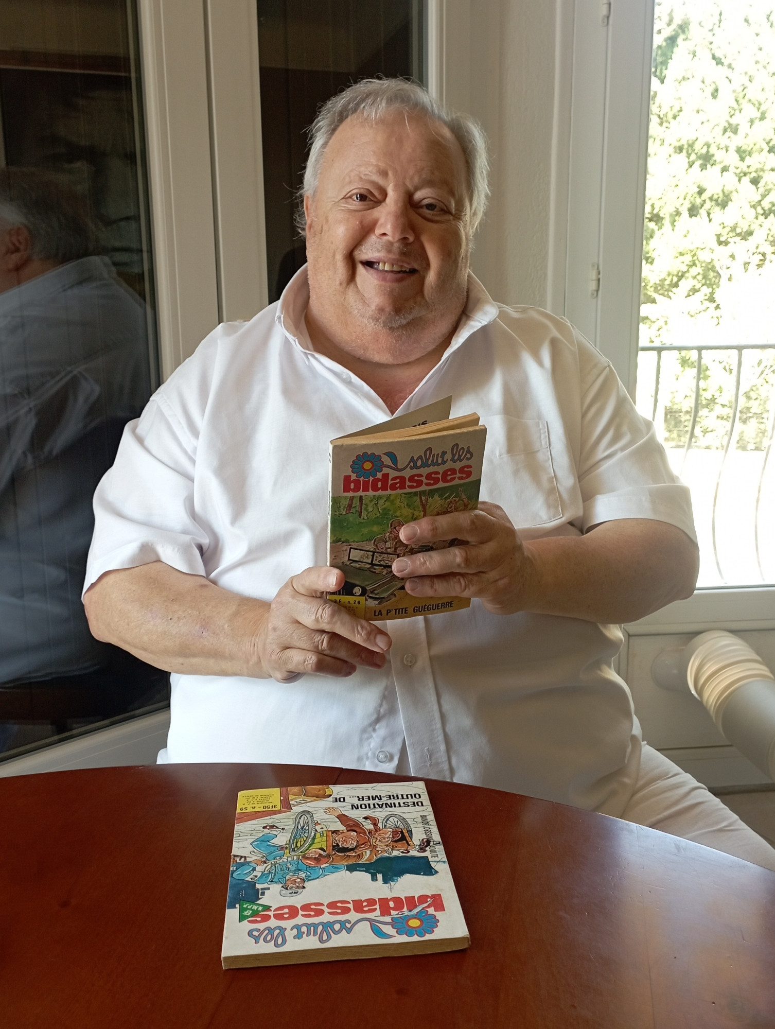 Photo de l'acteur Eddy Jabès, souriant et tenant en main une BD "Salut les bidasses".