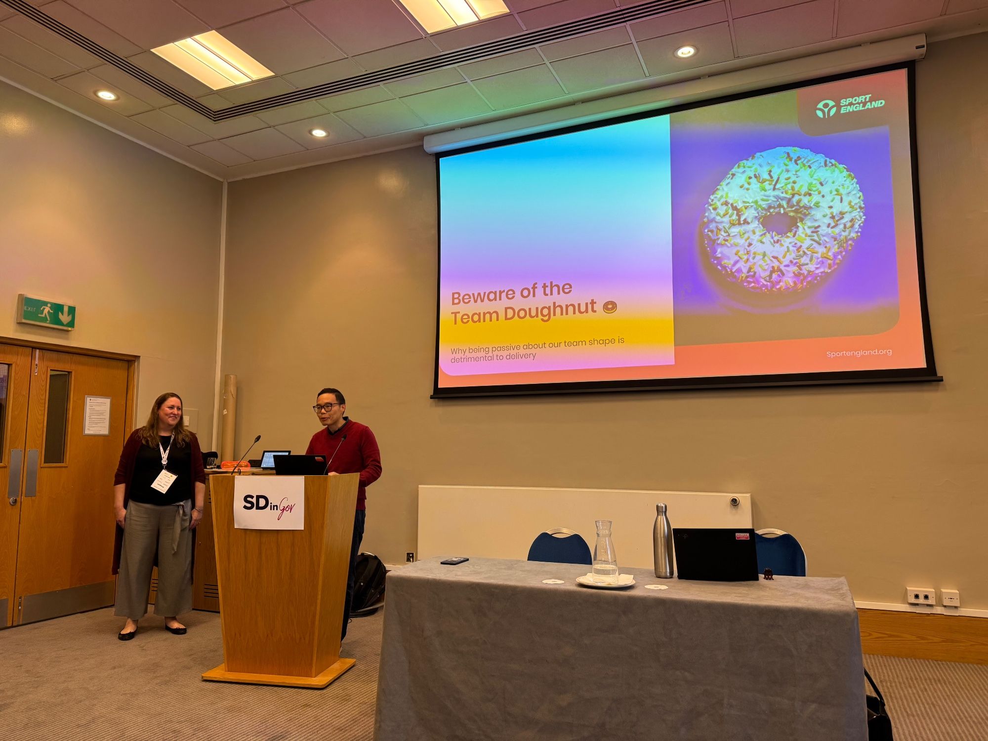 photograph of Cassie and Simon presenting in front of a big screen showing the headline slide