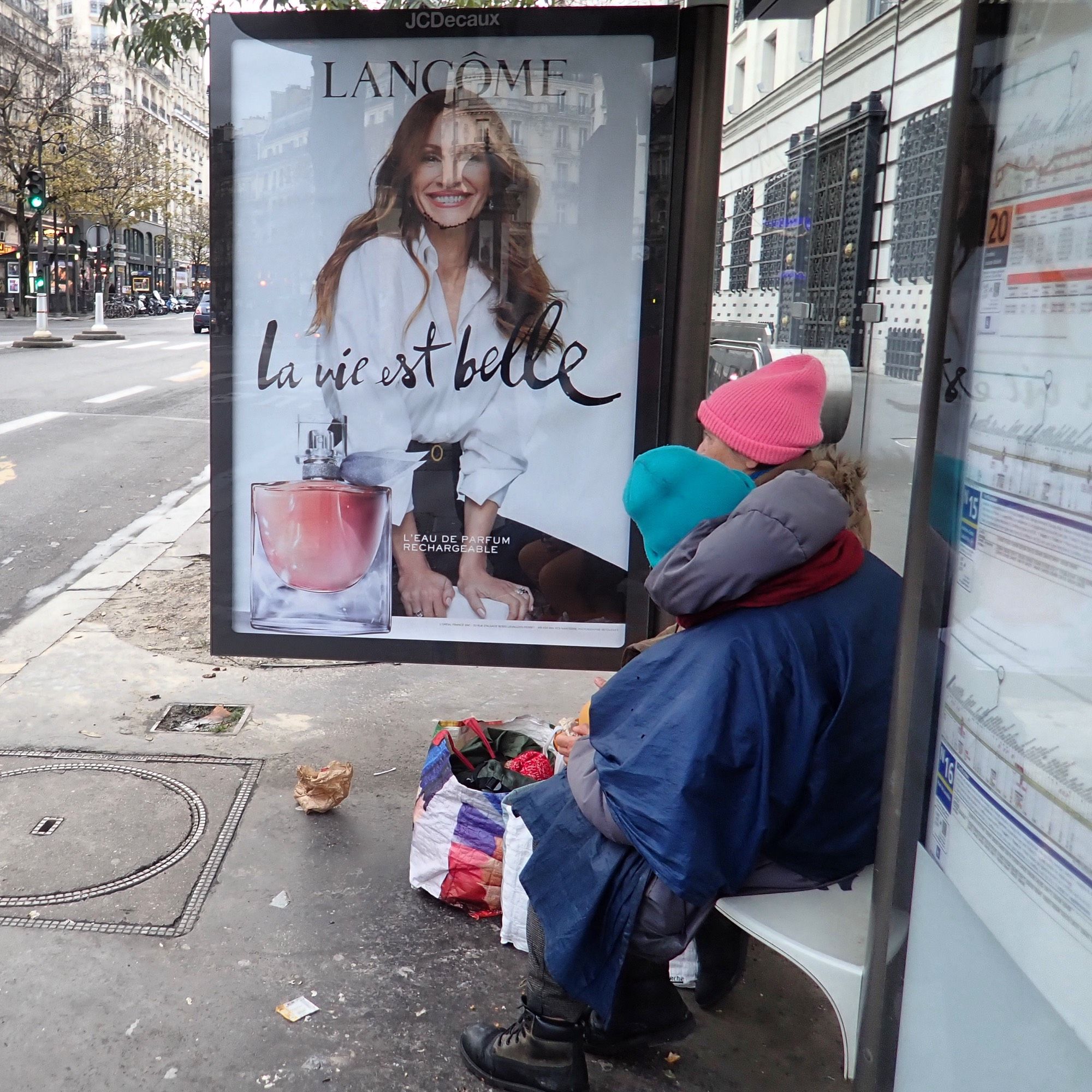 Deux femmes qui « vivent » dans la rue assises dans un arrêt de bus devant une publicité de Lancôme qui explique que « La vie est belle » avec une éclatante Julia Roberts.