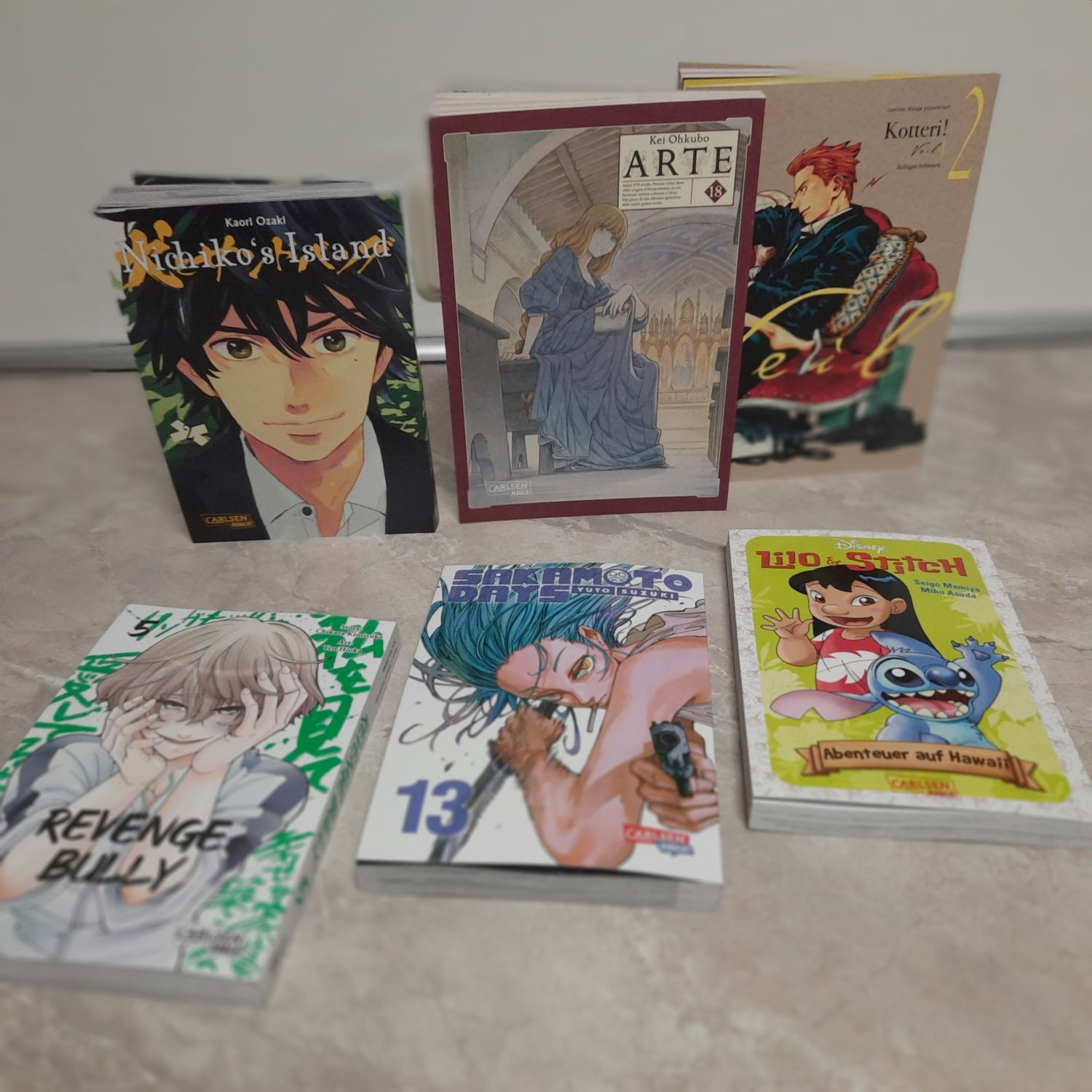 A collection of six manga and graphic novels are displayed on a table. The books are arranged in two rows. The top row features "Nichiko's Island" by Kaori Ozaki, "Arte" by Kei Ohkubo and "Koteri!" Vol. 2. The bottom row features "Revenge Bully" Vol. 5, "Sakamoto Days" Vol. 13 and "Lilo & Stitch: Abenteuer auf Hawai" by Seigo Miyamiya and Miho Asada. The books are all in German and feature a variety of genres, including romance, drama, action, and comedy. The covers of the books are colorful and eye-catching, and they are sure to appeal to fans of manga and graphic novels.