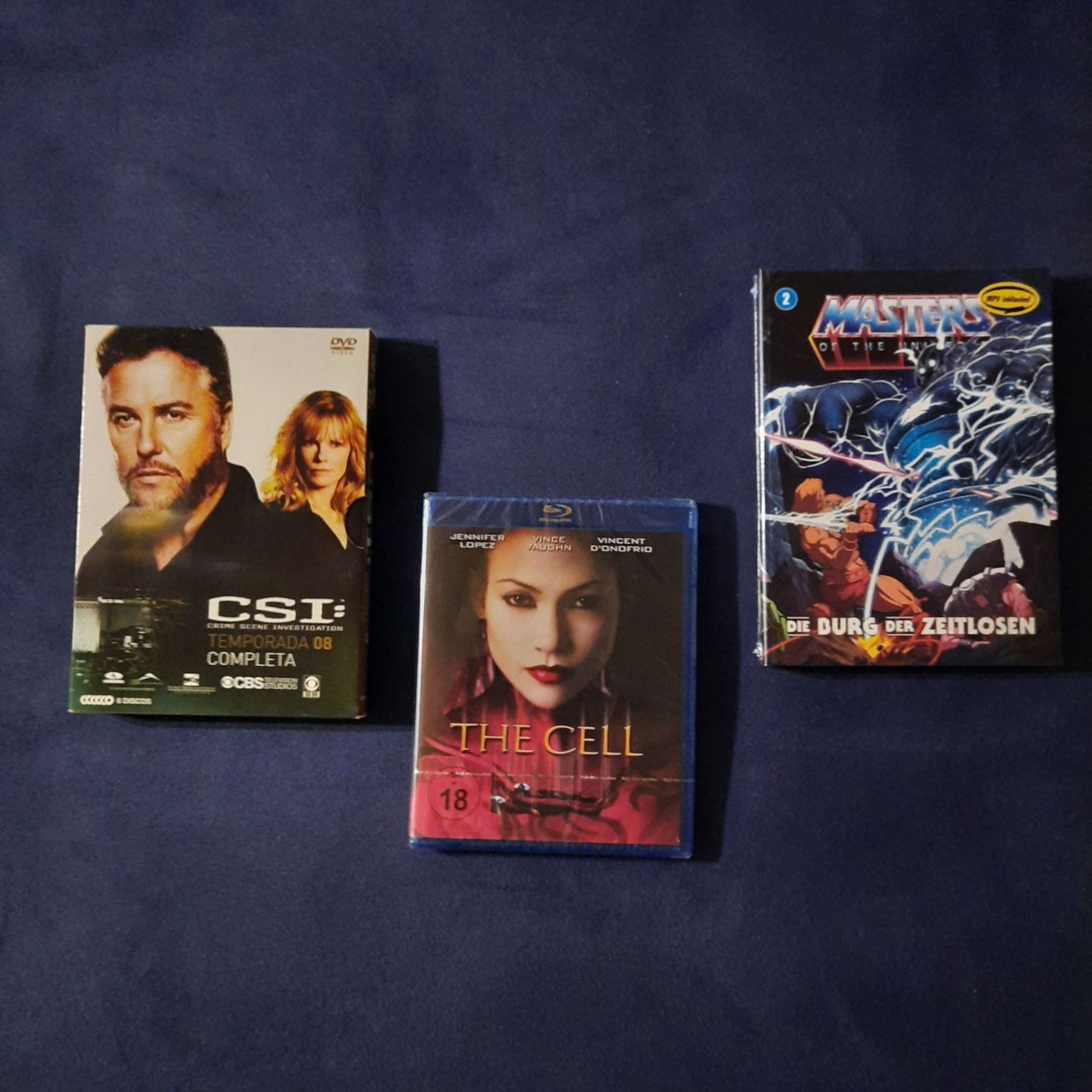 The image shows three DVD cases on a dark blue surface. The case on the left is for the TV show "CSI: Crime Scene Investigation" and features a man with dark hair and a serious expression. The middle case is for the movie "The Cell" and features a woman with dark hair and red lipstick looking directly at the camera. The case on the right is for the movie "Masters of the Universe" and features a muscular figure in a comic book style. The image is a good representation of a collection of DVDs.