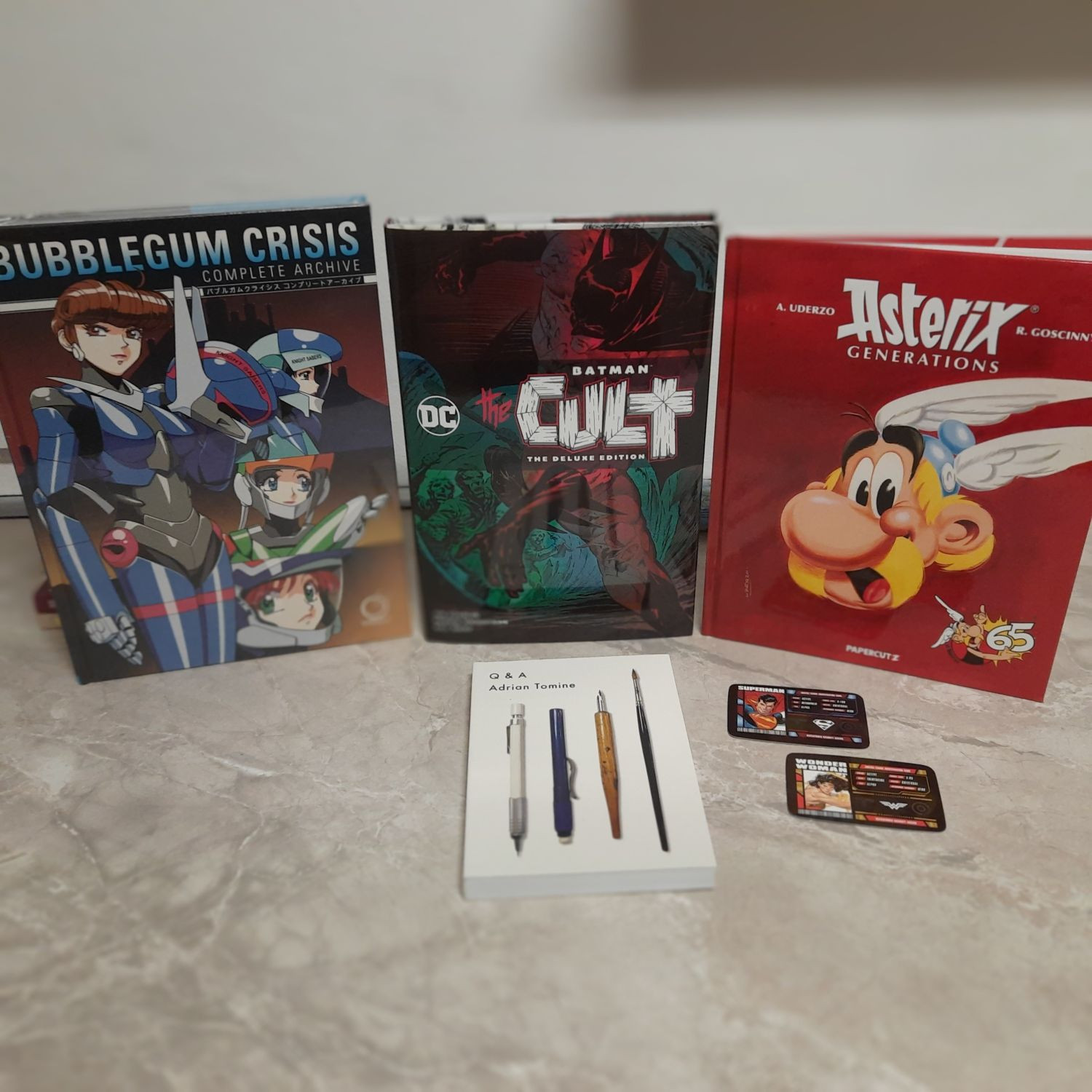 A collection of graphic novels and comics are displayed on a marble surface. The books are "Bubblegum Crisis Complete Archive," "Batman: The Cult Deluxe Edition," and "Asterix: Generations." There are also two trading cards featuring Superman and Wonder Woman, and two pens and a pencil. The image captures a casual and eclectic collection of popular culture items.