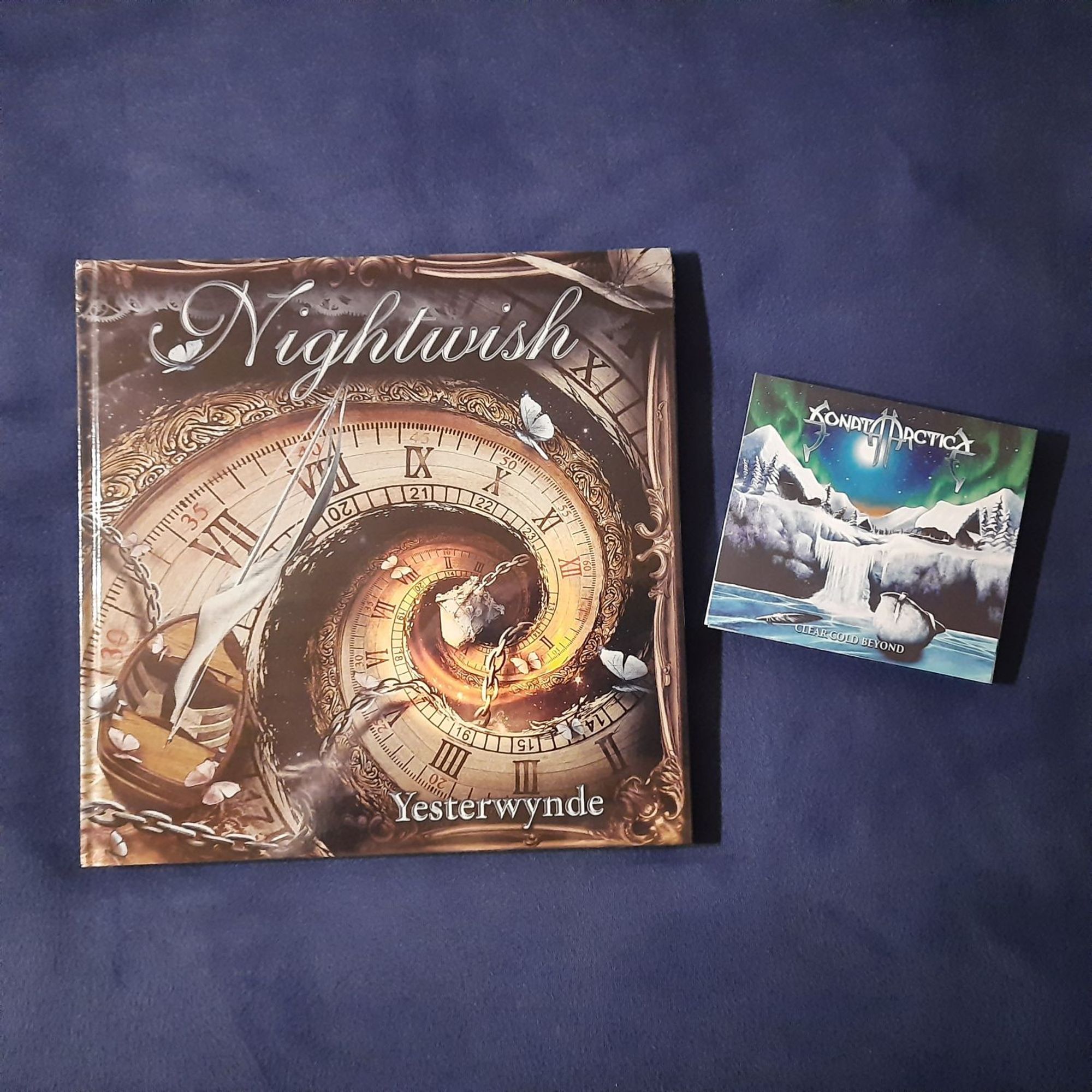 The image shows two CDs on a blue surface. The CD on the left is a Nightwish album titled "Yesterwylde". The cover art features a clock face with Roman numerals and a chain, surrounded by butterflies and other decorative elements. The CD on the right is a Sonata Arctica album titled "Clear Cold Beyond". The cover art features a snowy landscape with a waterfall and a full moon in the sky. The image is well-lit and the CDs are in focus.