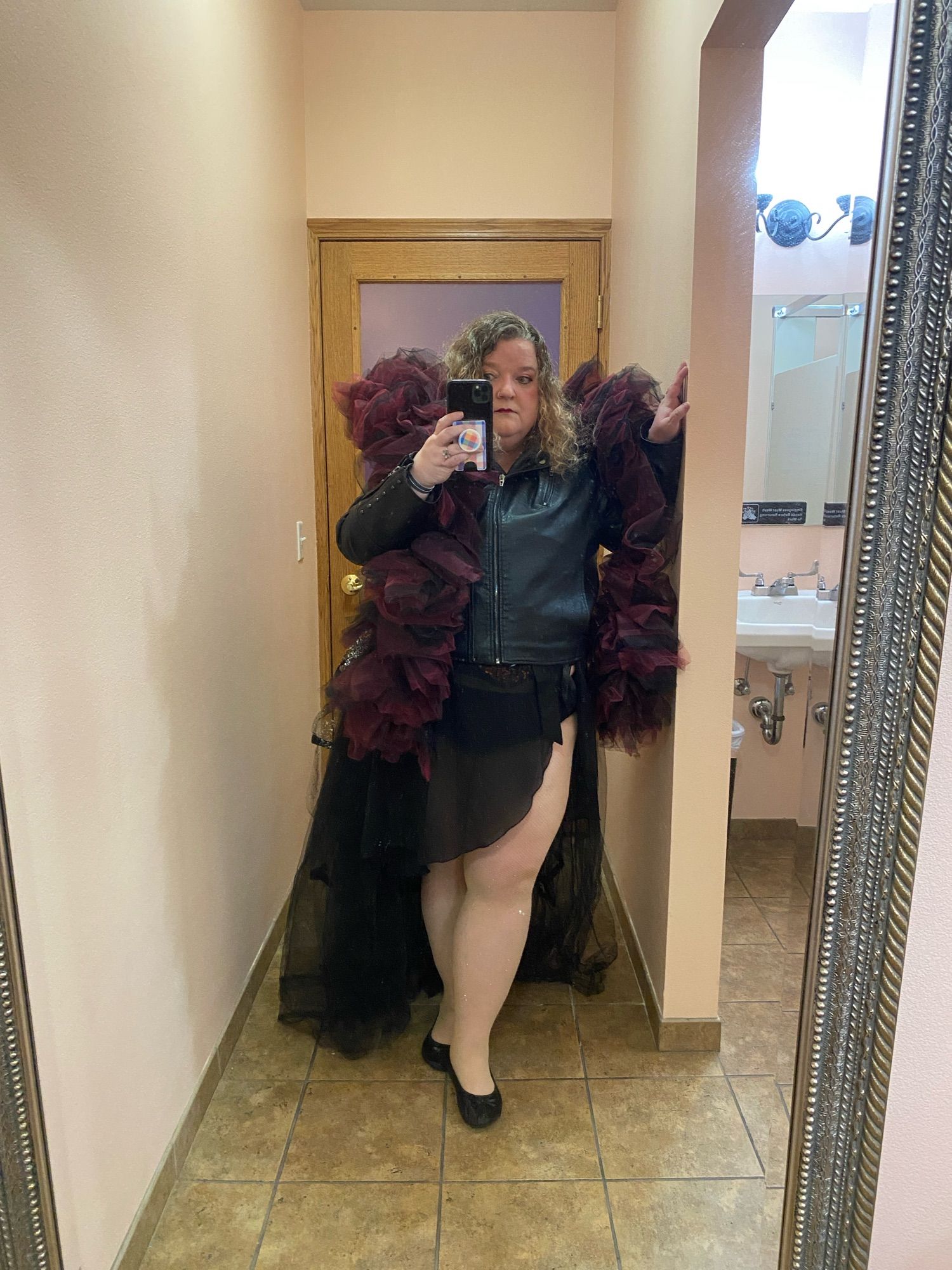 Me standing and posing in a bathroom mirror wearing a black leather jacket, black skirt, long black tulle bustle, and a black & burgundy tulle boa around my shoulders