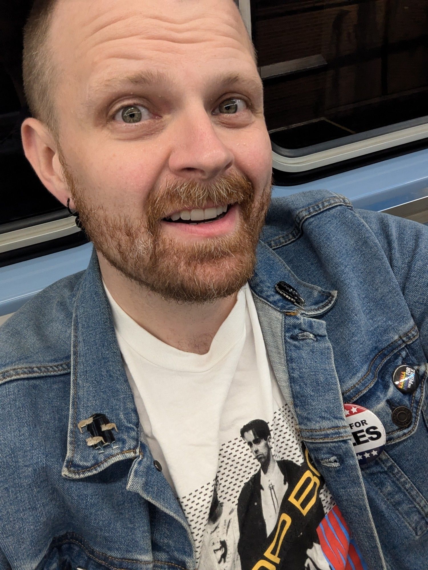 A bearded man smiles. He's wearing a denim jacket over a Pet Shop Boys "West End Girls" tee shirt. On the jacket are pins of Edward Gorey (and his cats) and the Bride of Frankenstein plus buttons for IDLES and The People for Kamala Harris.