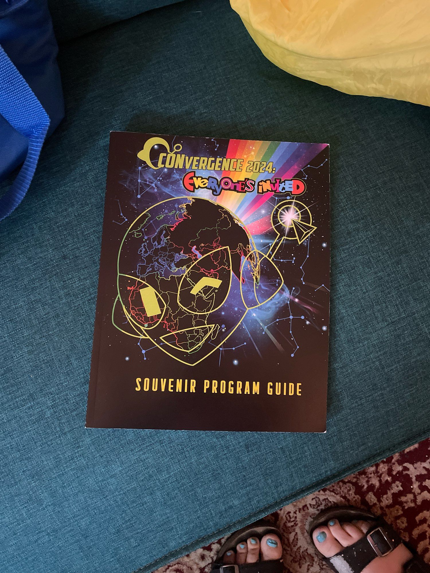 The CONvergence 2024 souvenir program guide resting on a couch. A black and rainbow paperback with a gynoid robot (Connie) on the cover