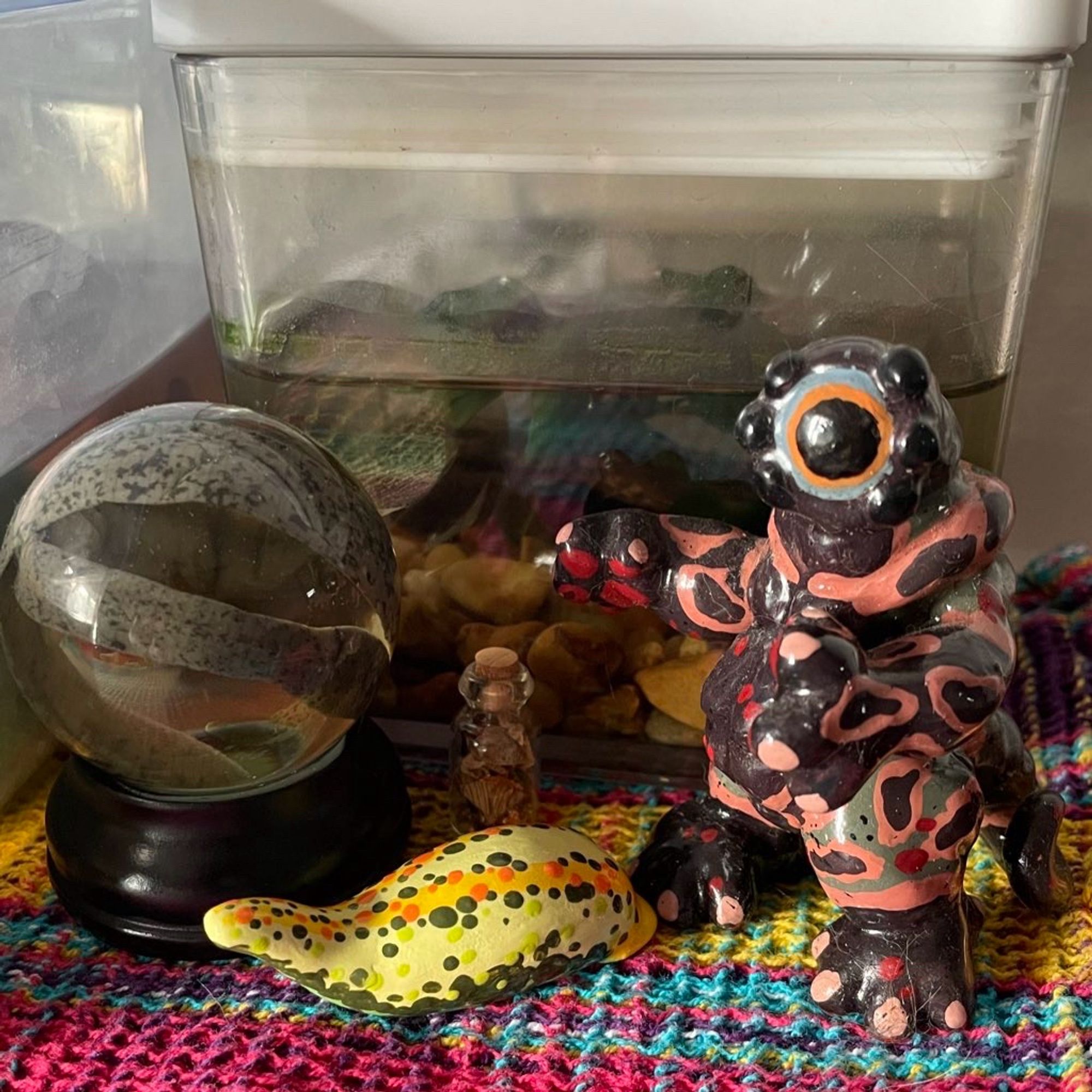 A leech sculpture, two preserved leeches in a glass orb, a small glass bottle with dried leeches and an anthro leech cyclops character stand on a rainbow placemat in front of a leech tank