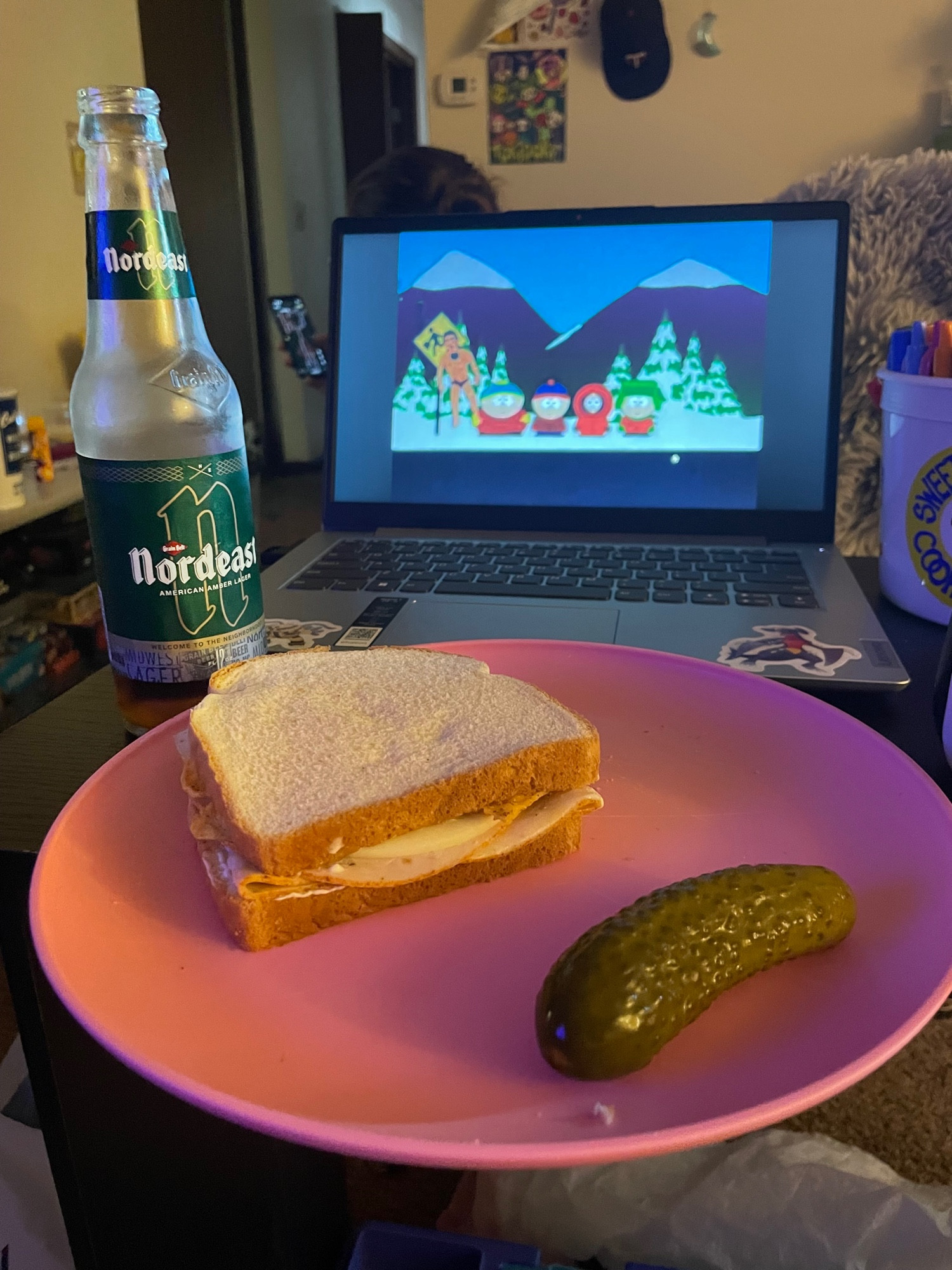 beer, a chicken and cheese sandwich with mayo and chips, and a pickle, with south park playing off a laptop