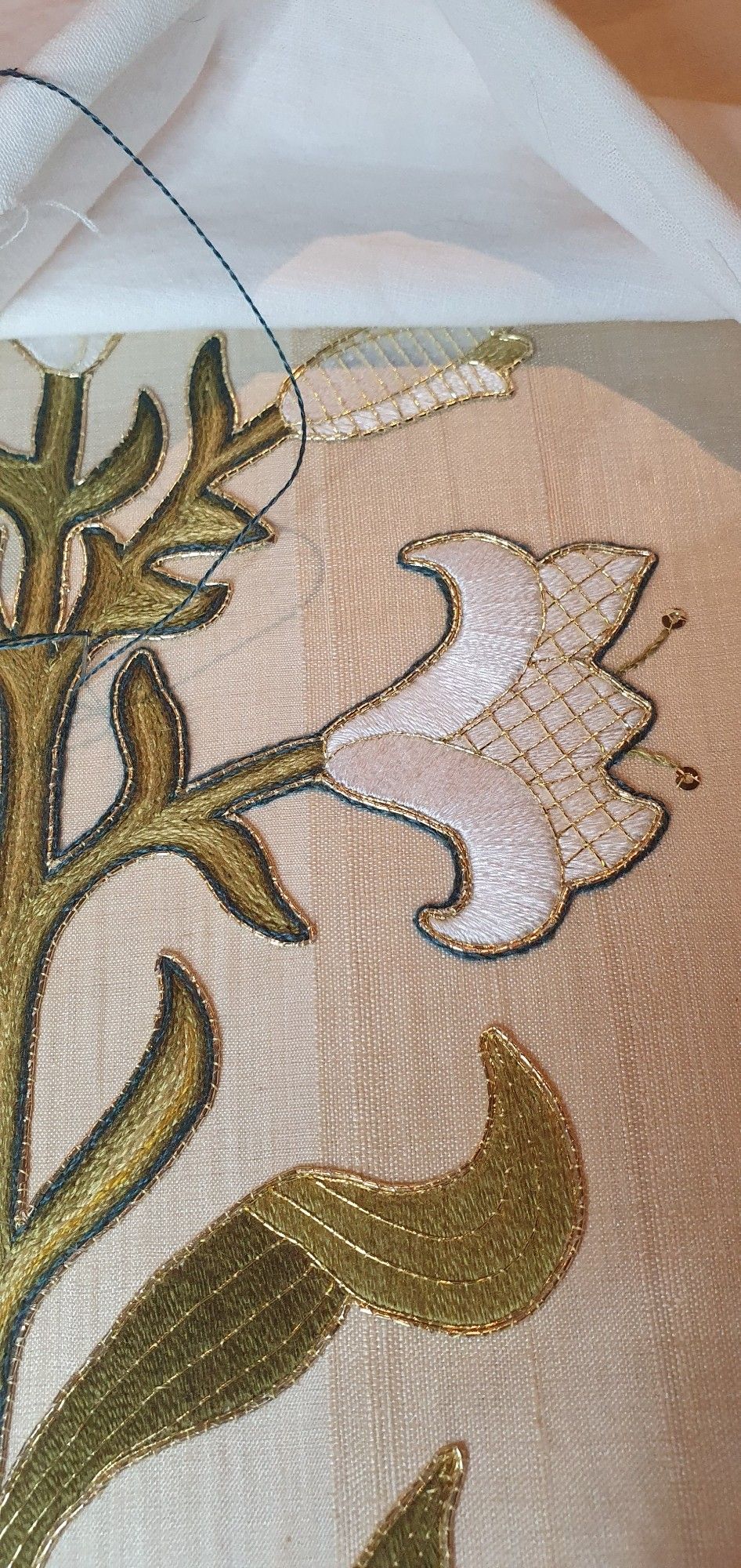 Close up of an embroidered white lily with green leaves, worked in silk and gold