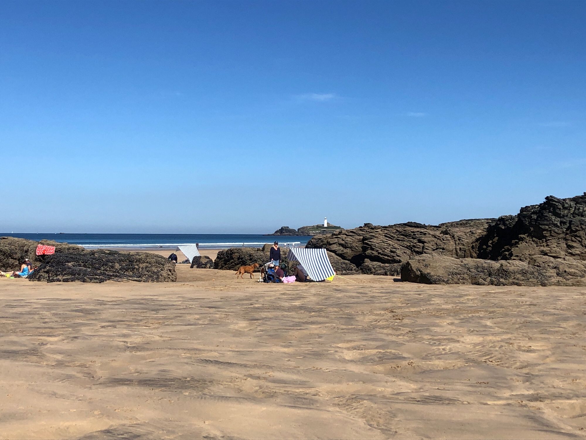 A sunny, Sandy beach with Rockpools, a lighthouse on an island and two matching blue and white sunshade / windbreak contraptions on frames