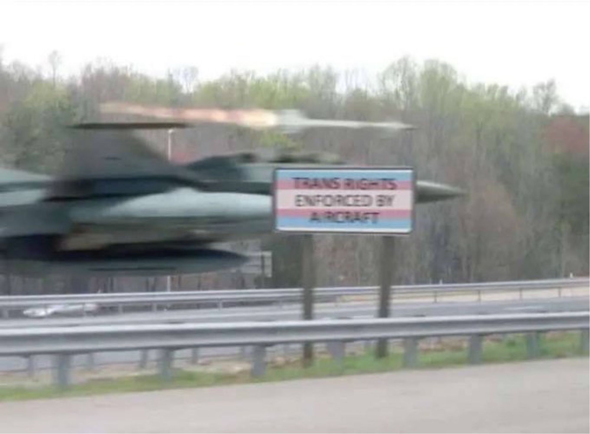 A jet fighter flying behind a sign that reads “Trans Rights Enforced By Aircraft” on a highway media . The sign has a trans flag background.