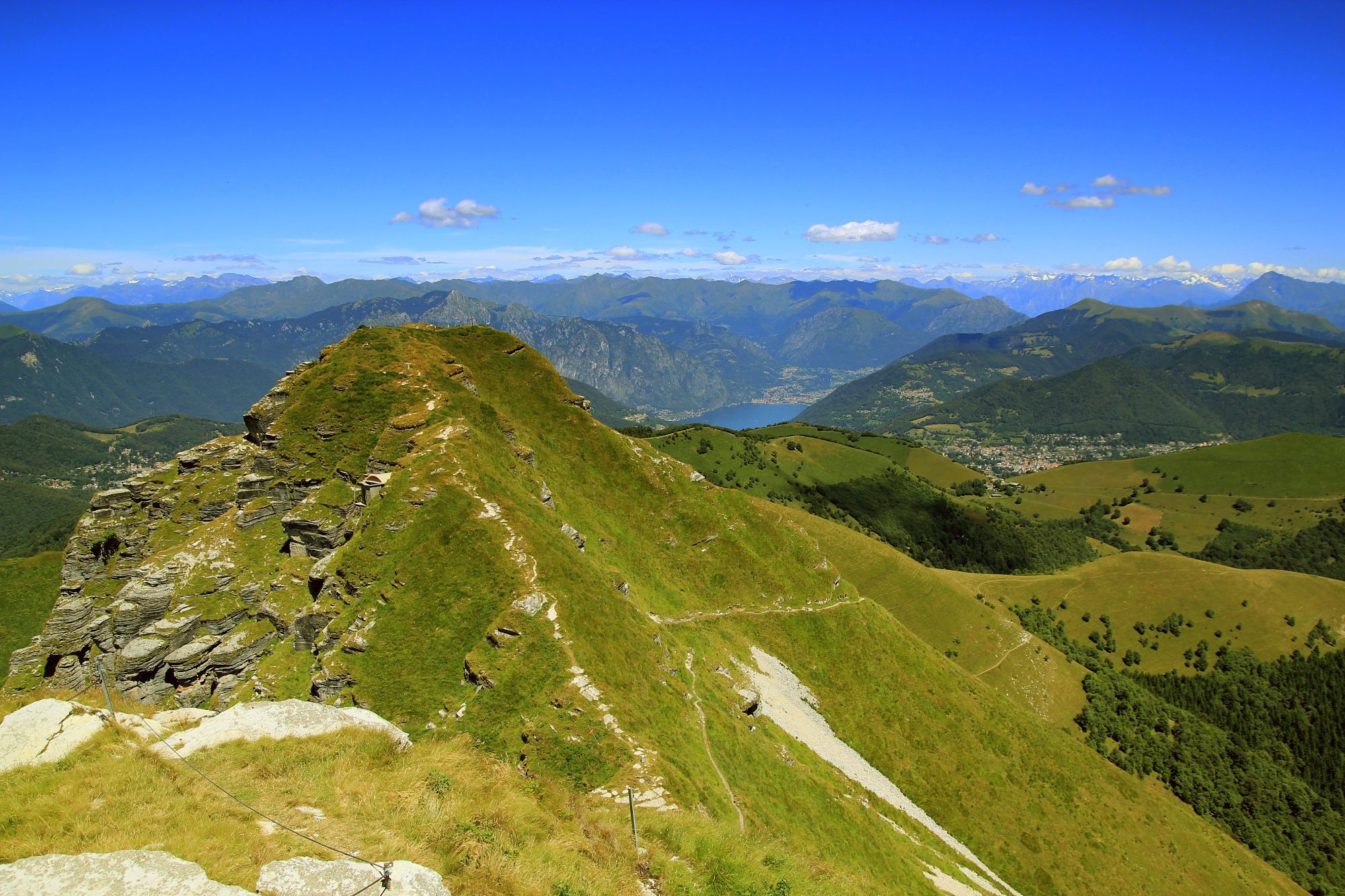 Monte Generoso - Ein Bergblick mit grünen Gipfeln und im Hintergrund, weit entfernt sind die Alpen zu erkennen. Der Himmel ist weitestgehend blau.