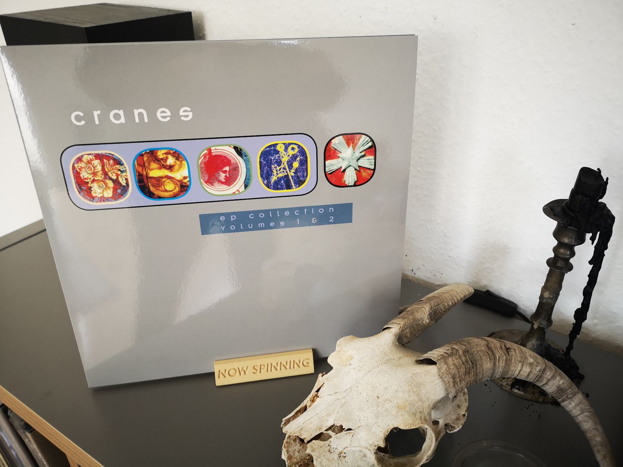 Ein graues LP Cover mit fünf kleineren Abbildungen, welche ebenfalls LP Cover zeigen. Dazu die Aufschrift 'Cranes - EP Collection 1 & 2'

Davor ist ein Ziegenschädel und ein Kerzenständer zu sehen.